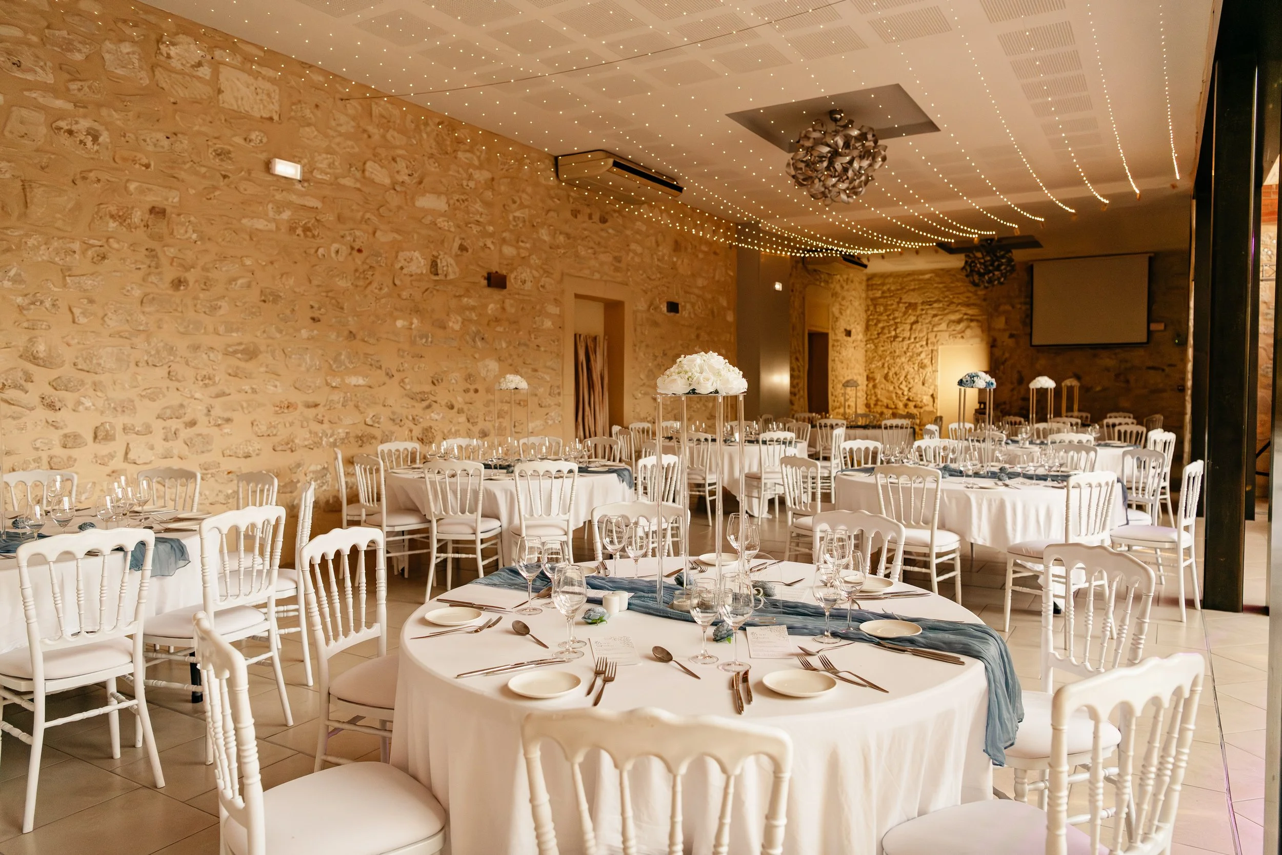 Salle de réception décorée pour un événement avec des tables rondes avec nappes blanches, chaises blanches, centre de table floral blanc sur une table, guirlandes lumineuses au plafond, murs en pierre, grande projection vide à l'arrière, ambiance élé