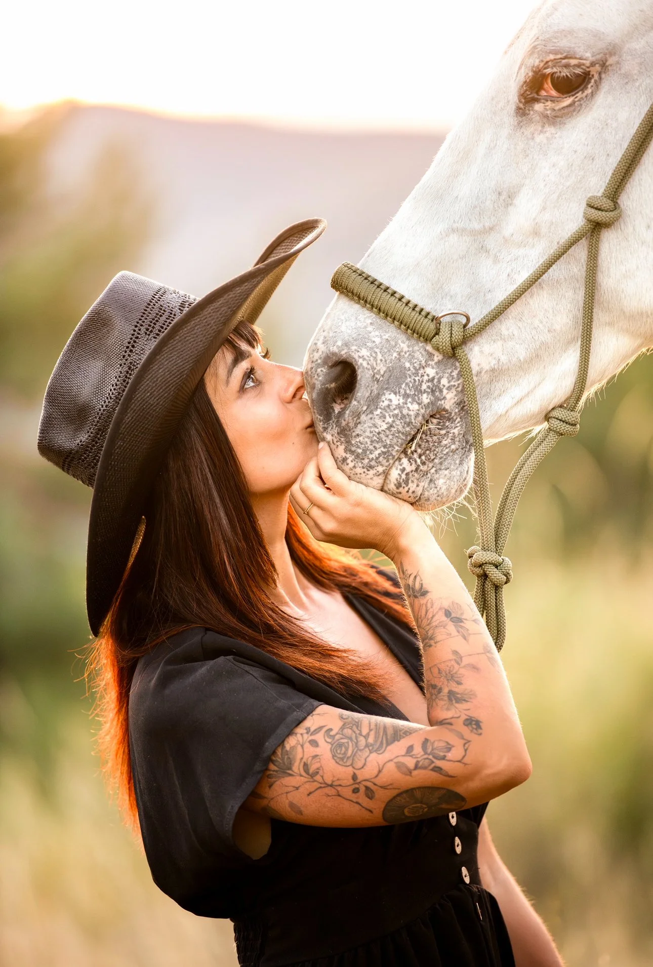 Une femme tatouée portant un chapeau noir embrasse un cheval blanc dans un champ lors du coucher de soleil.