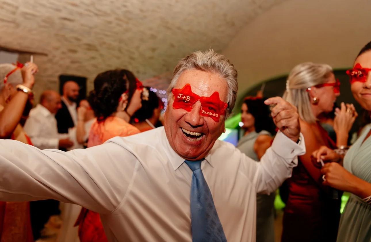 Un homme souriant portant des lunettes en forme d'étoile rouge avec des initiales et une date, lors d'une fête, entouré d'autres personnes habillées pour une célébration.