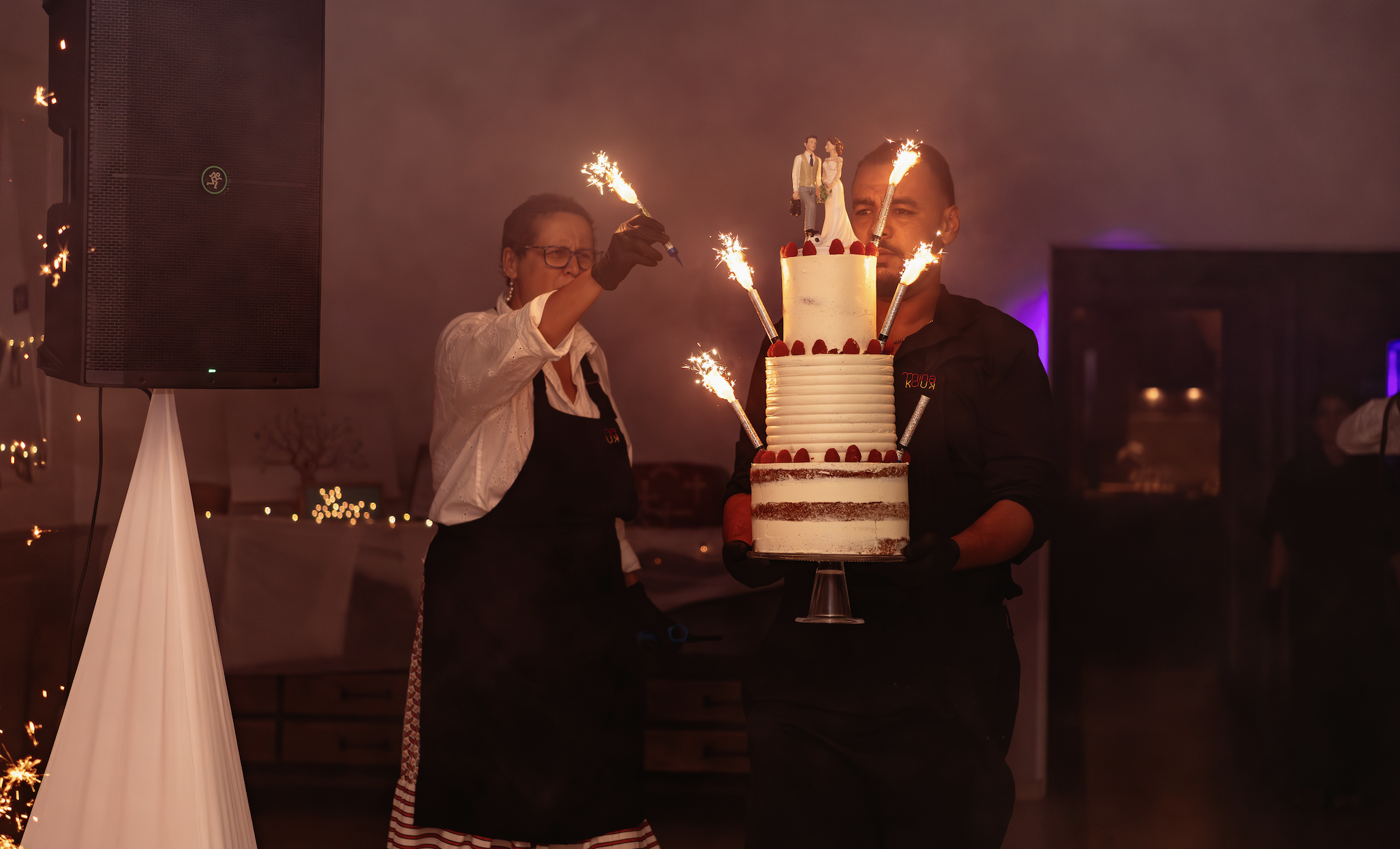 Un homme tient un gâteau d'anniversaire à plusieurs niveaux décoré de rubans rouges et de figurines, avec des étincelles de feux d'artifice.