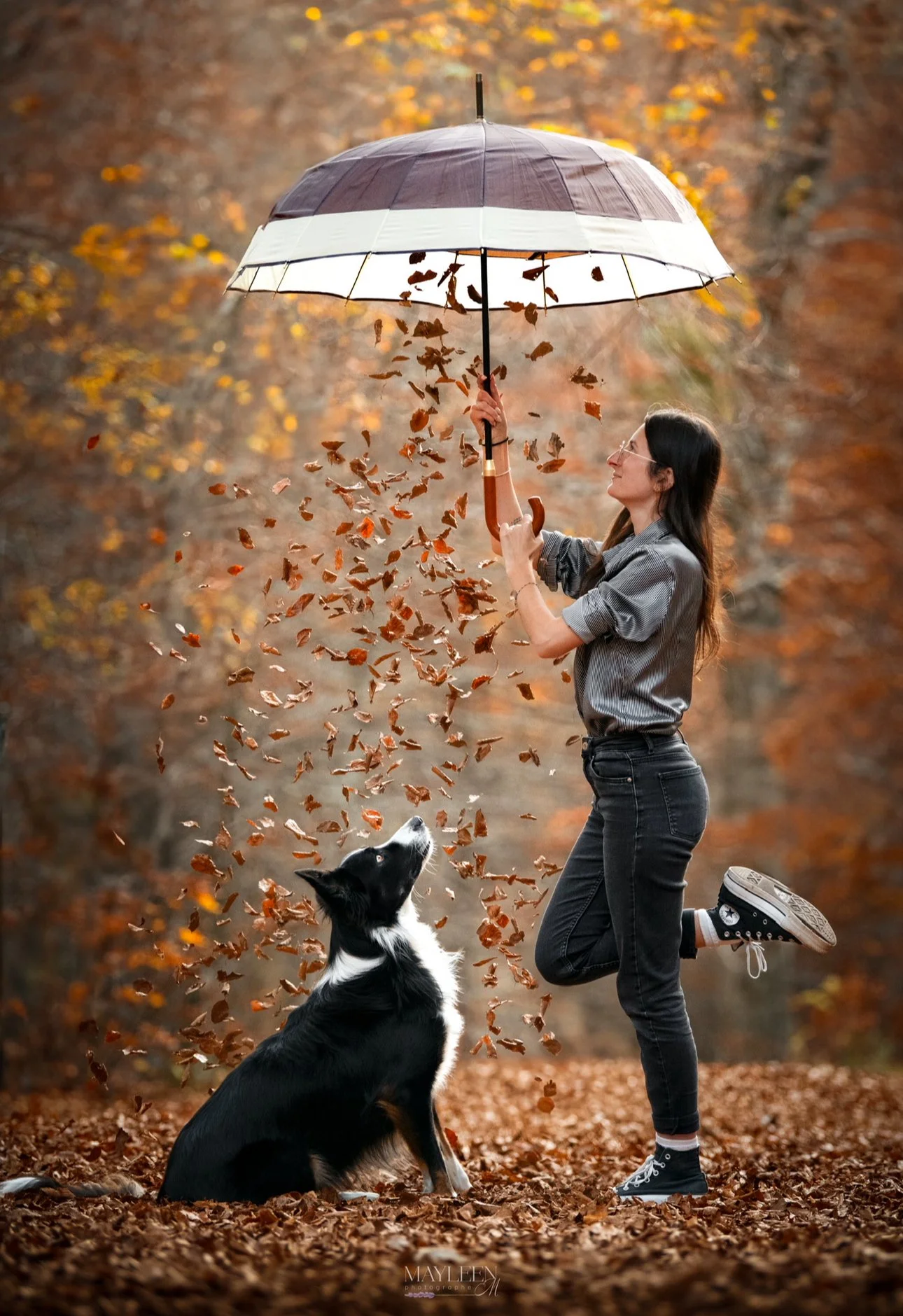 Une femme avec un parapluie et un chien dans un parc en automne, tous deux entourés de feuilles tombantes.