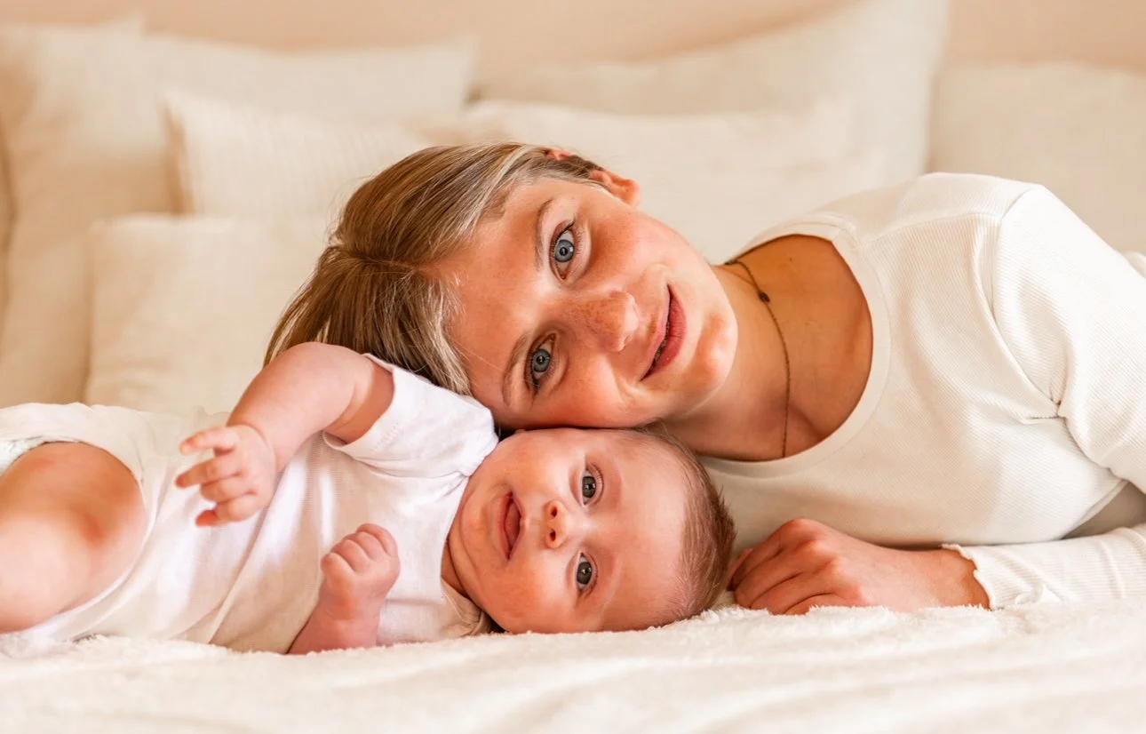Une femme et un bébé allongés sur un lit, souriant à la caméra, dans un décor beige et doux.