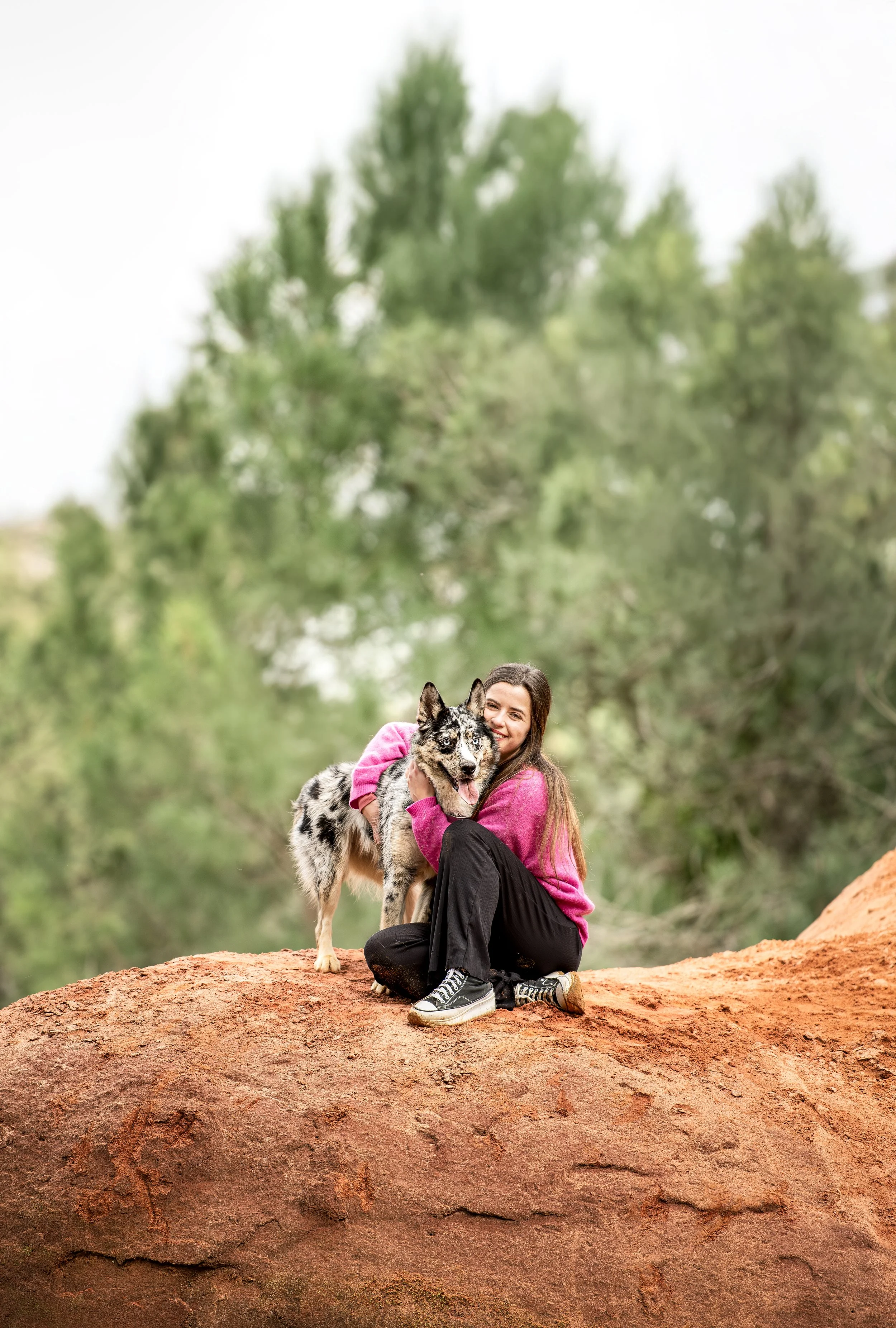 Une femme souriante en pull rose et pantalon noir assise sur une roche, avec un chien de race husky aux couleurs variées, dans un environnement naturel avec des arbres en arrière-plan.