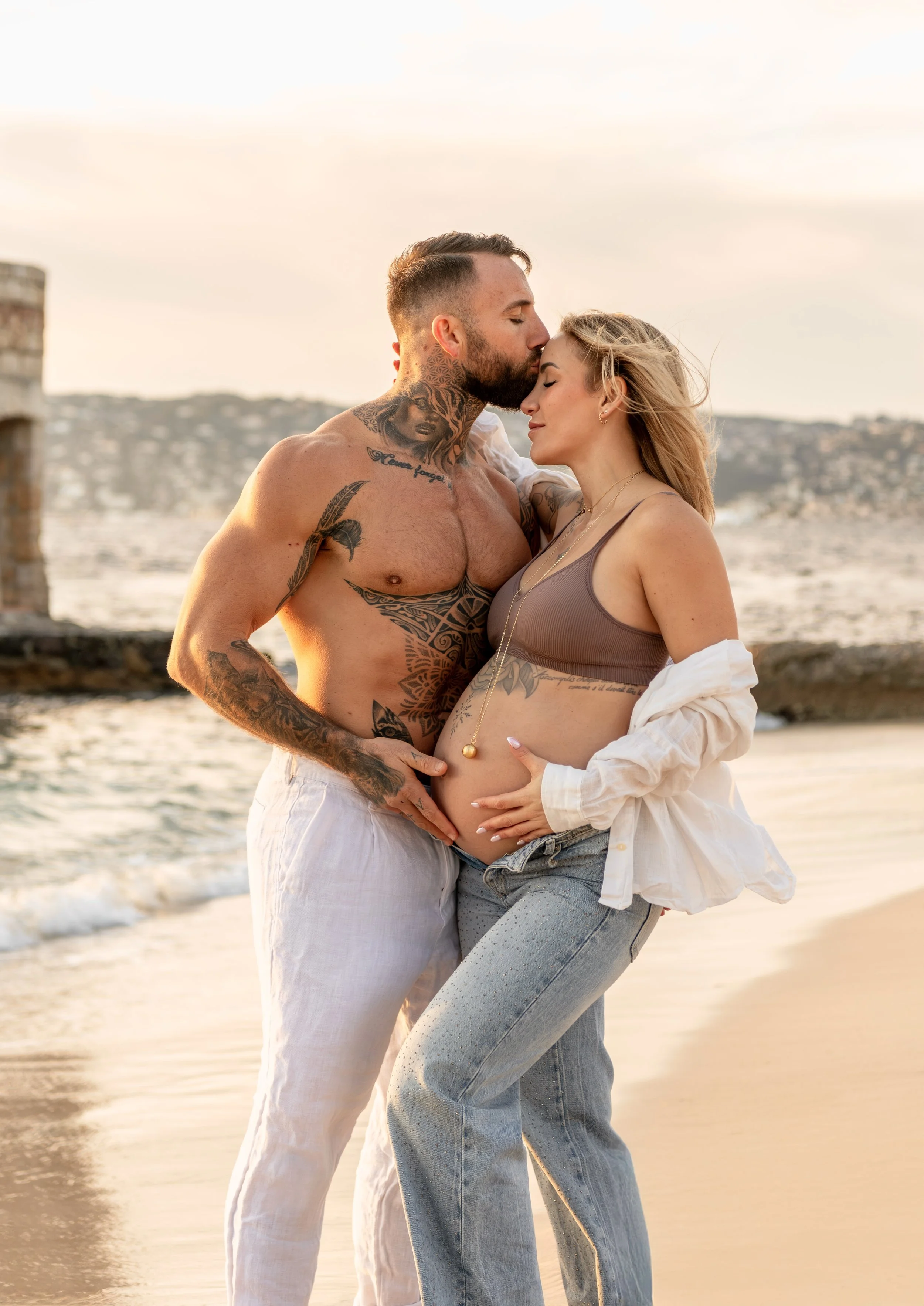 Un couple debout sur la plage au coucher du soleil, la femme enceinte, lui embrassant le front, mains sur son ventre, le fond d'une mer, des bâtiments en arrière-plan.