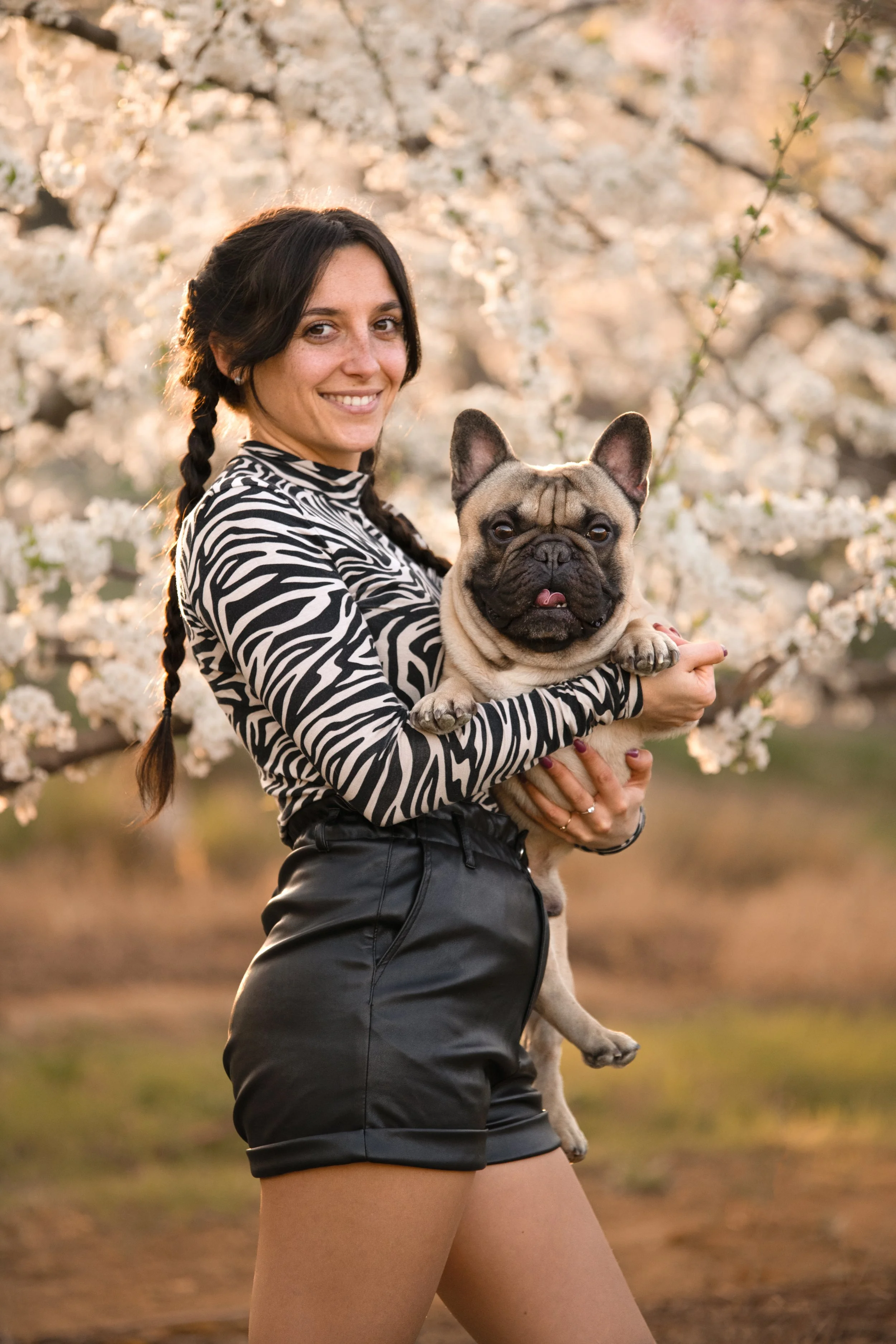 Une femme souriante avec une robe zébrée noire et blanche, tenant un bouledogue français dans un jardin en fleurs au printemps.