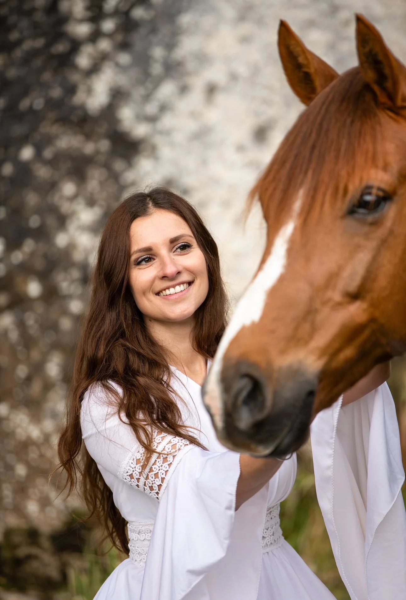 Une femme souriante en robe blanche caresse un cheval brun avec une tache blanche sur le visage, en extérieur.