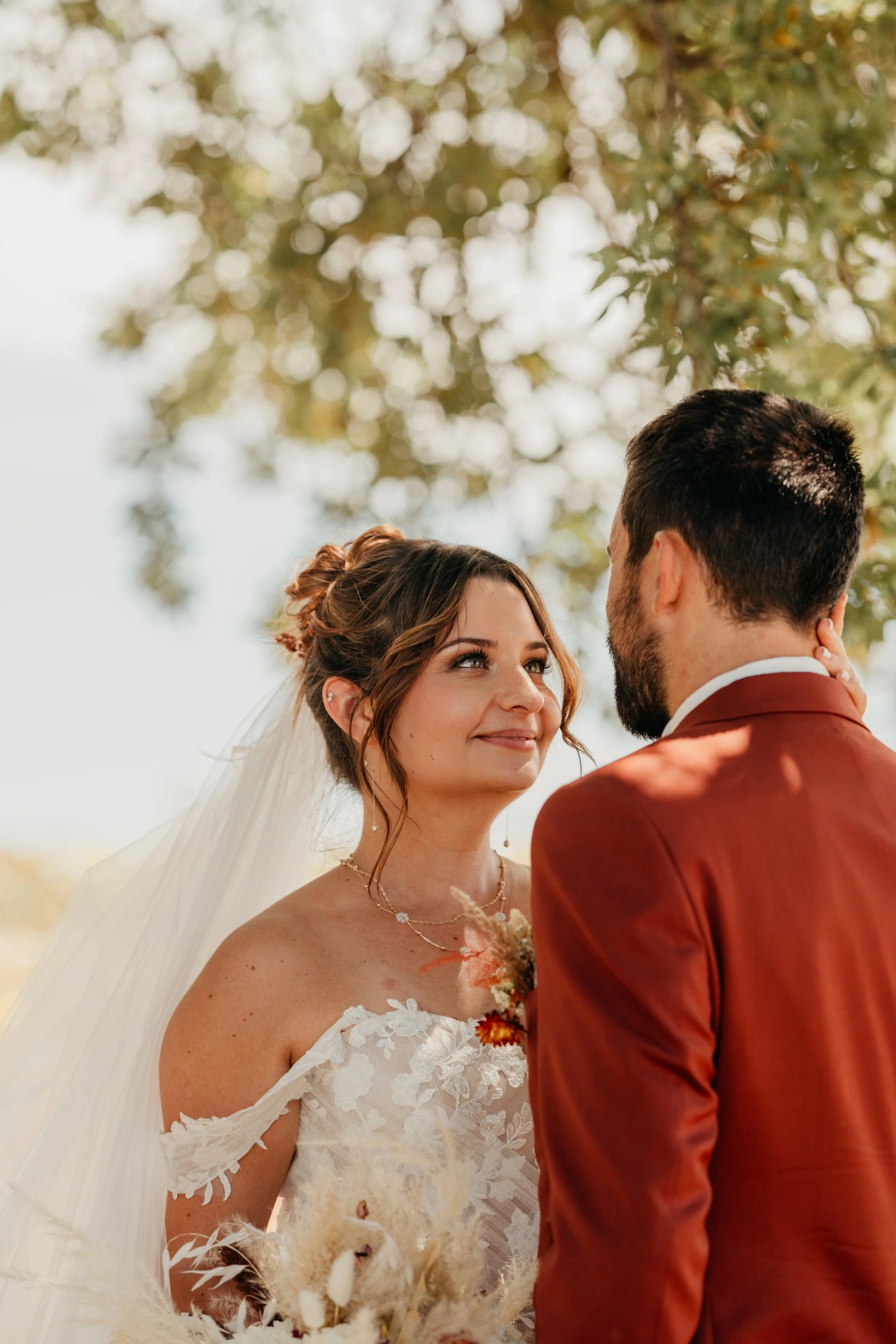 Un couple lors de leur mariage, la femme en robe blanche avec un voile et un bouquet, l'homme en costume marron, se regardant tendrement sous un arbre.