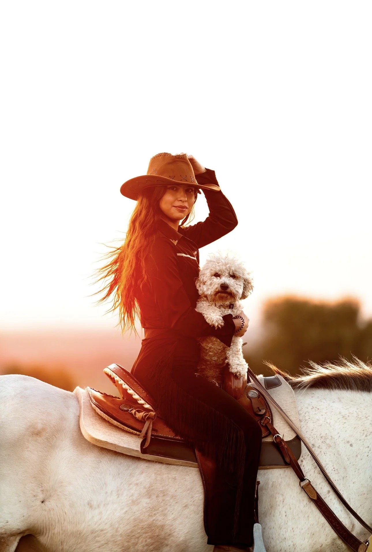 Une femme en habits de cowgirl sur un cheval blanc, tenant un chien blanc, lors d'un coucher de soleil.
