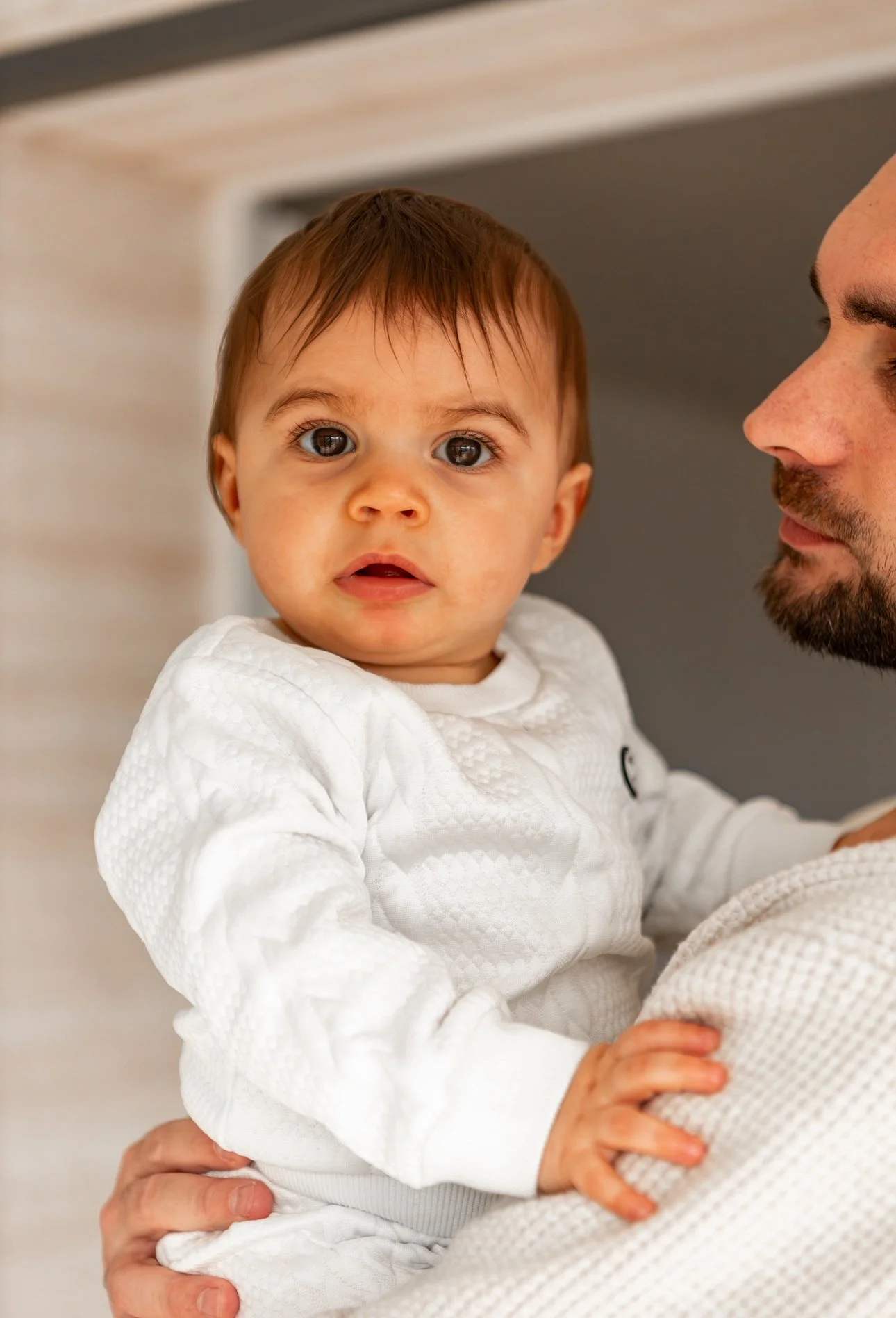 Un bébé avec des cheveux bruns, portant un vêtement blanc, est tenu par un homme dont le visage n'est pas complètement visible pendant une séance photo famille en intérieur 