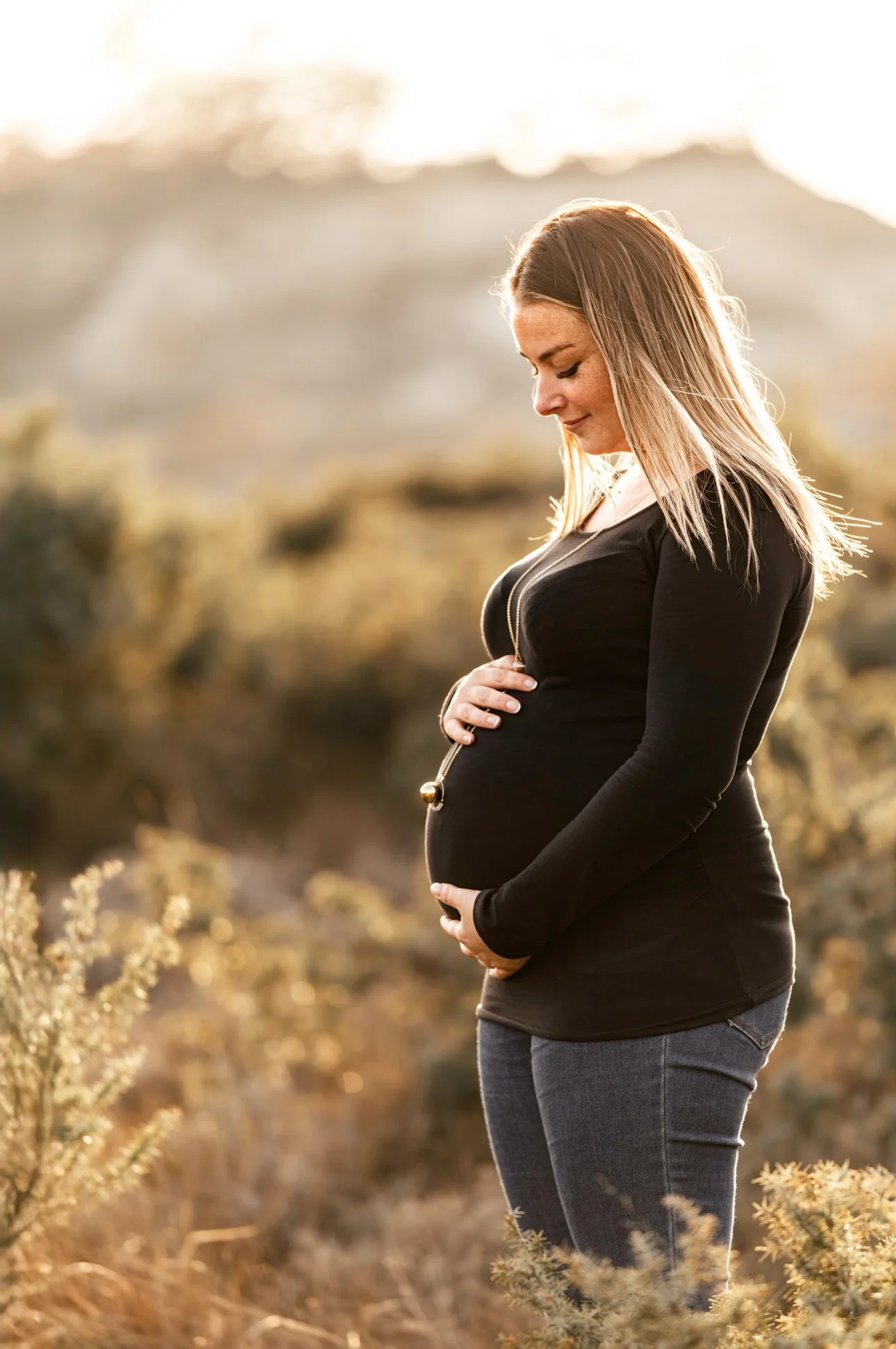 Une femme enceinte, portant un haut noir, se tient dans un champ naturel lors d'un coucher de soleil, tenant son ventre.