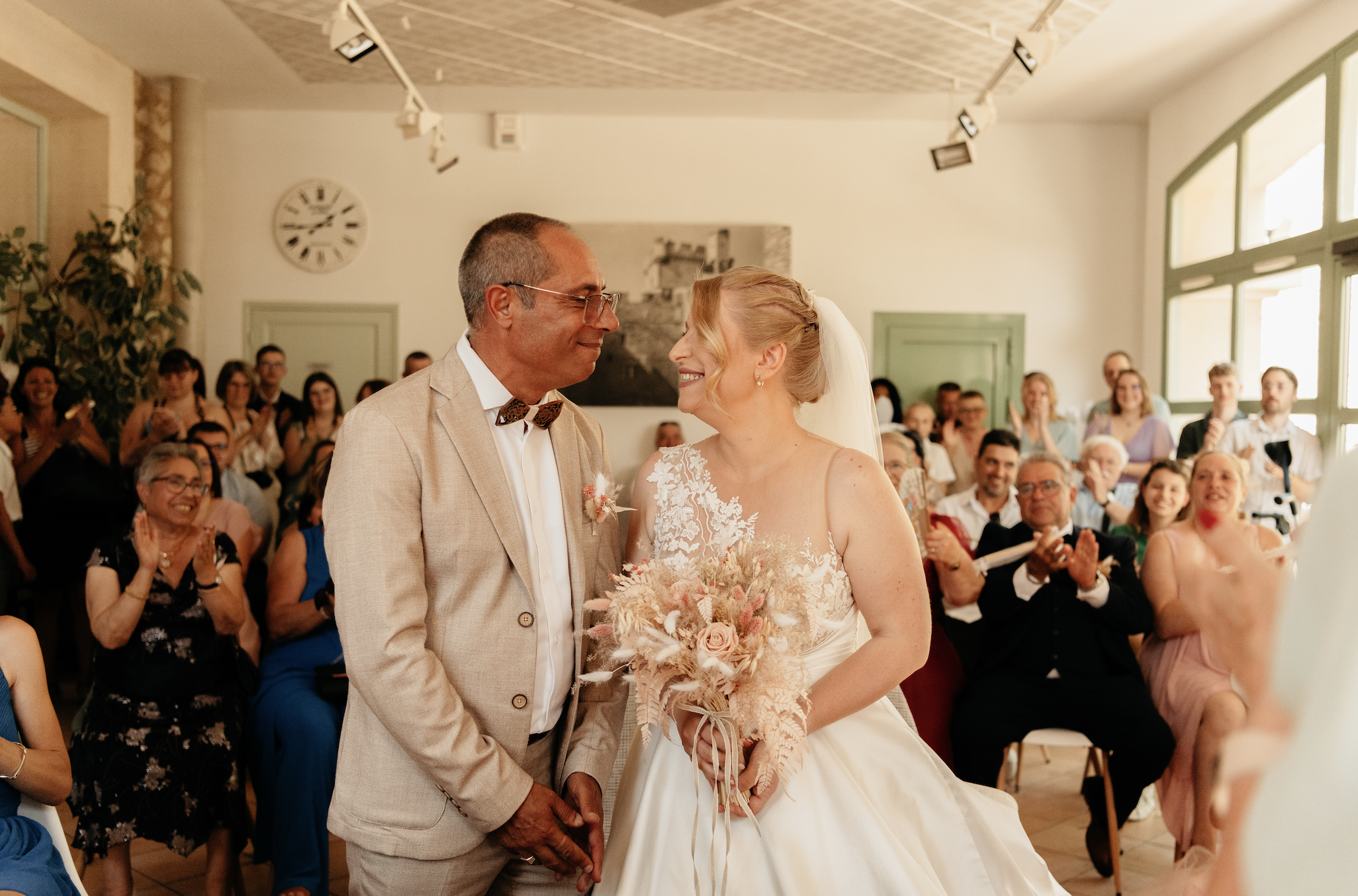 Un couple de mariés échangeant leur vœux lors de leur mariage entourés de invités dans une salle lumineuse.