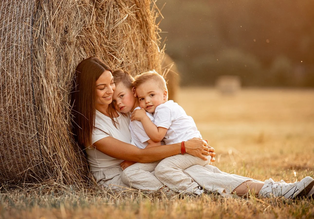Une femme et deux enfants assis près d'un tas de foin dans un champ lors d'un coucher de soleil.