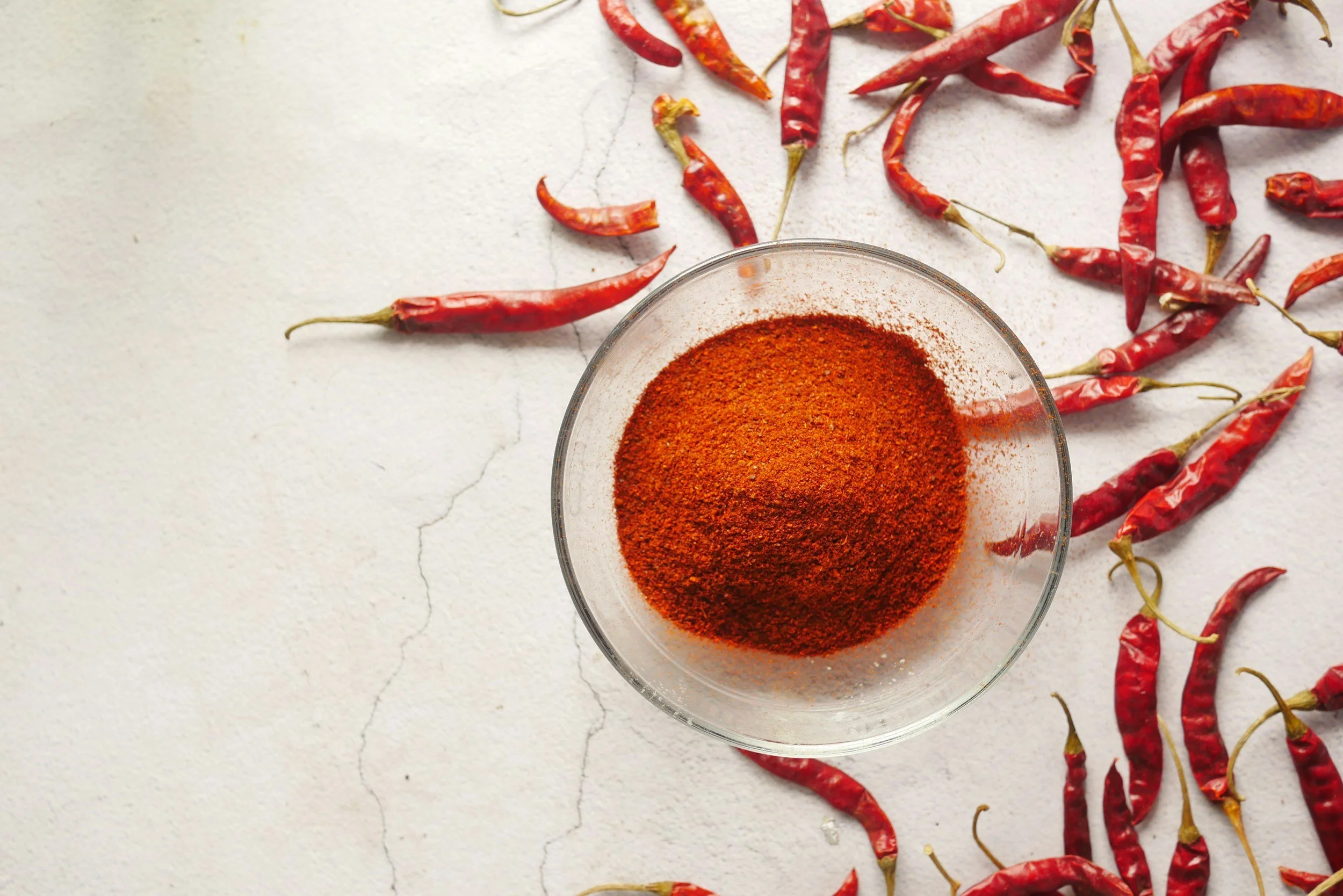 A glass bowl containing ground red chili powder surrounded by whole dried red chili peppers on a white textured surface.