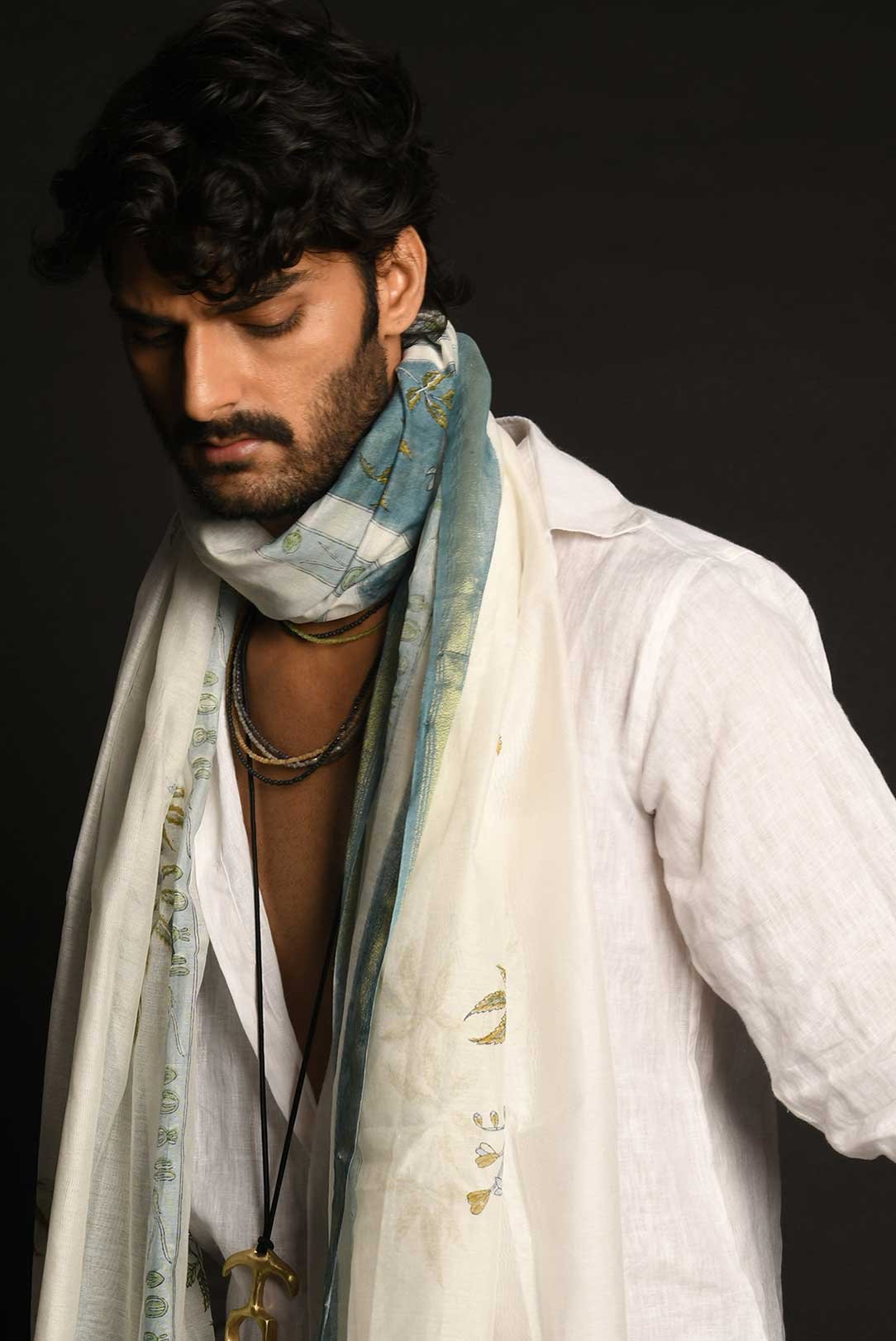 Man in a white shirt with a multicolored scarf and necklaces against a dark background.