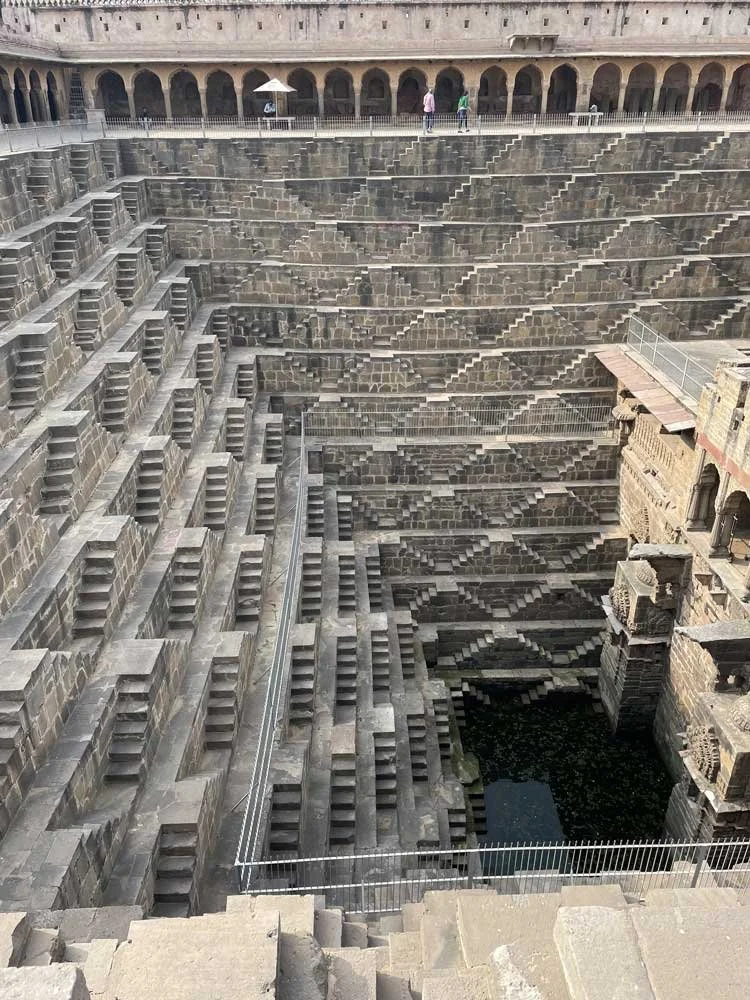 Ancient stepwell with symmetrical stone steps and railings, leading down to a pool of water at the bottom.