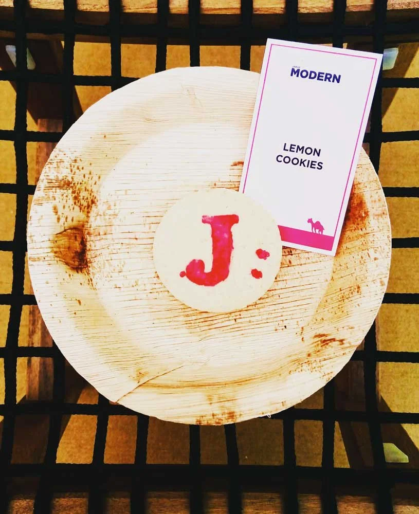 A round lemon cookie with a red letter 'J' on it is placed on a wooden plate. Behind the plate is a label that reads 'The Modern' and 'Lemon Cookies.' The items are set on a black grid surface.
