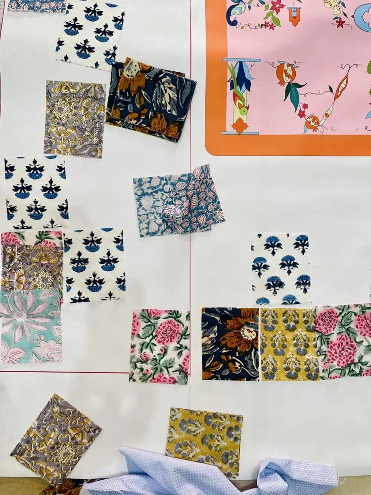 A display of various floral fabric swatches arranged on a white board with red lines and a pink decorative border. The swatches feature different floral patterns and colors, including blue, white, pink, and gold. A piece of blue fabric is draped at t