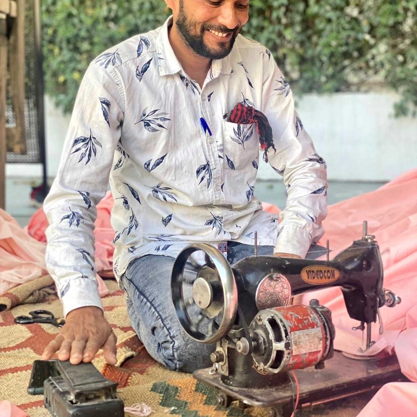 A man sitting on the floor smiling while using a vintage-style sewing machine with a foot pedal, surrounded by fabric and greenery in the background.