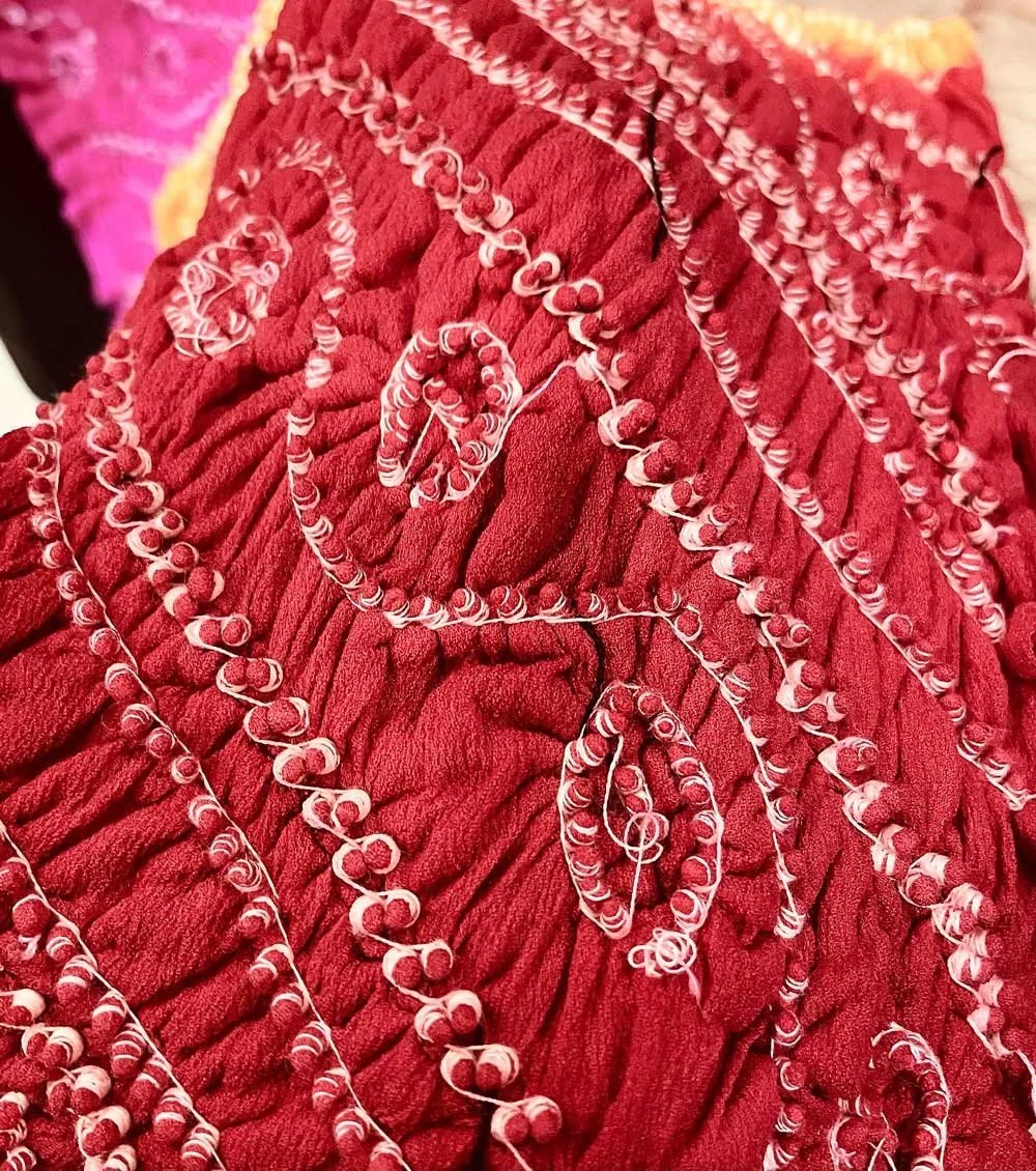 Close-up of red bandhani tie-dye fabric with intricate white patterns, featuring a crinkled texture.