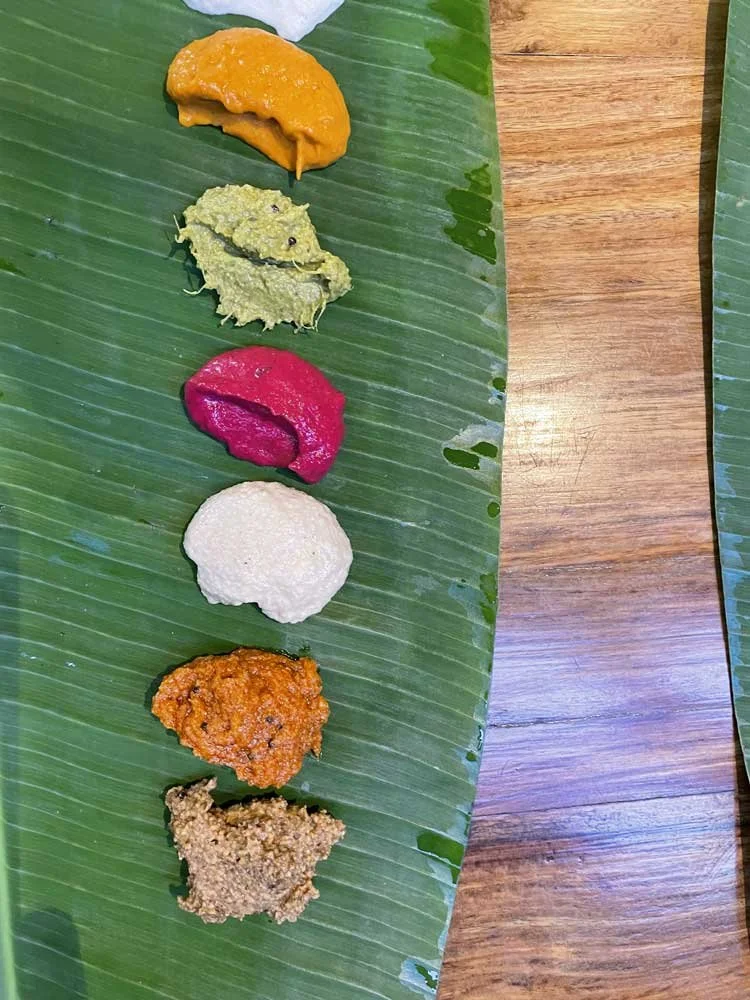 Various chutneys arranged in a line on a banana leaf on a wooden table.