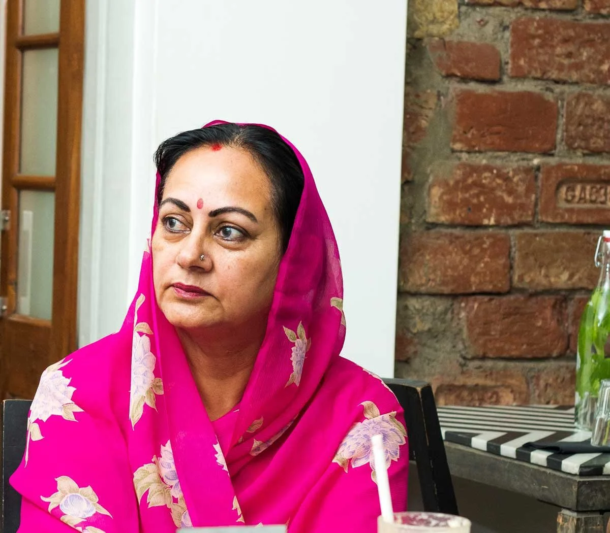 Maharini wearing a floral-patterned pink scarf with a red bindi on her forehead, sitting indoors next to a brick wall.