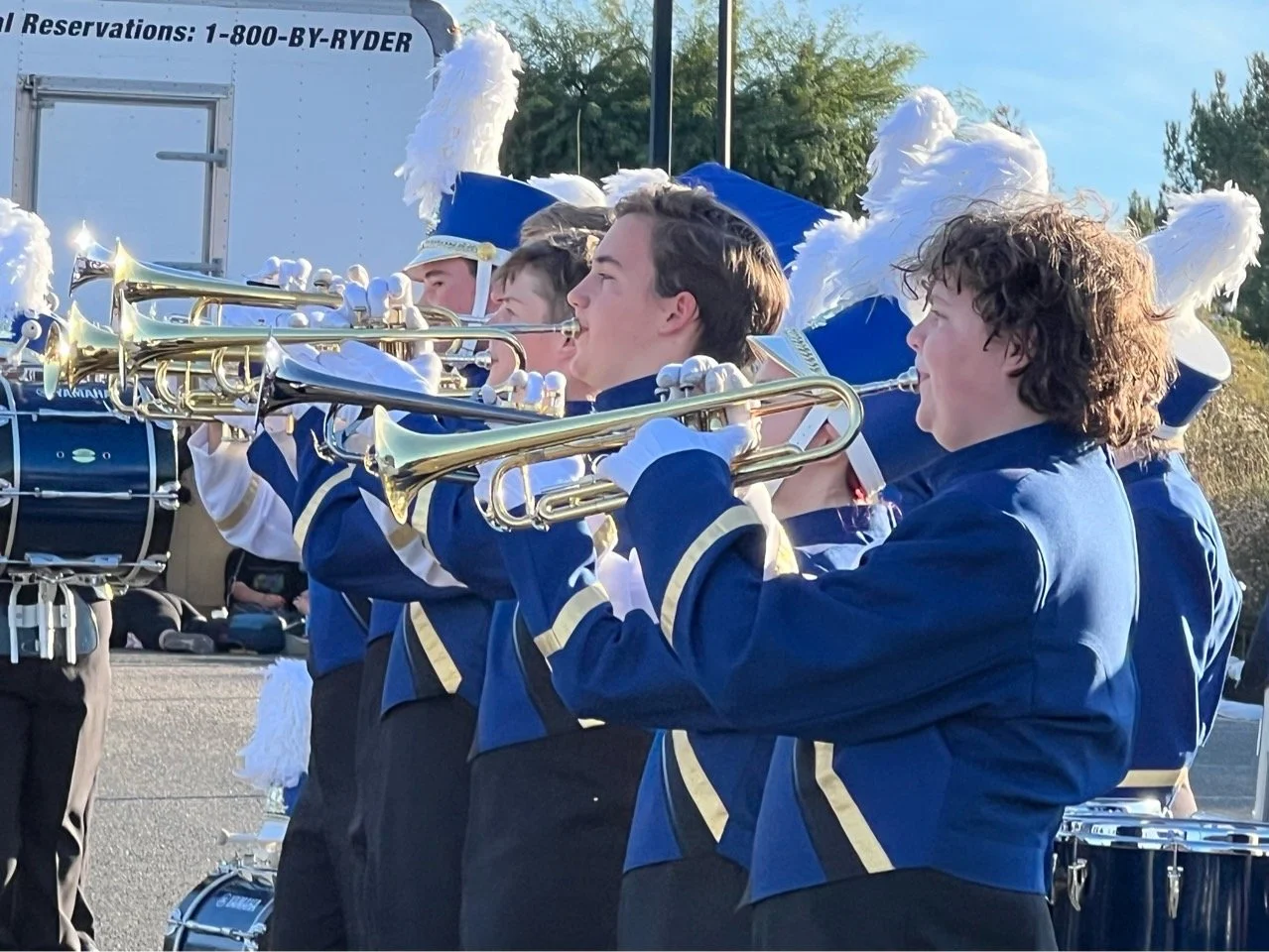 Young marching band members in blue uniforms with white feathered hats playing trumpets during a parade.