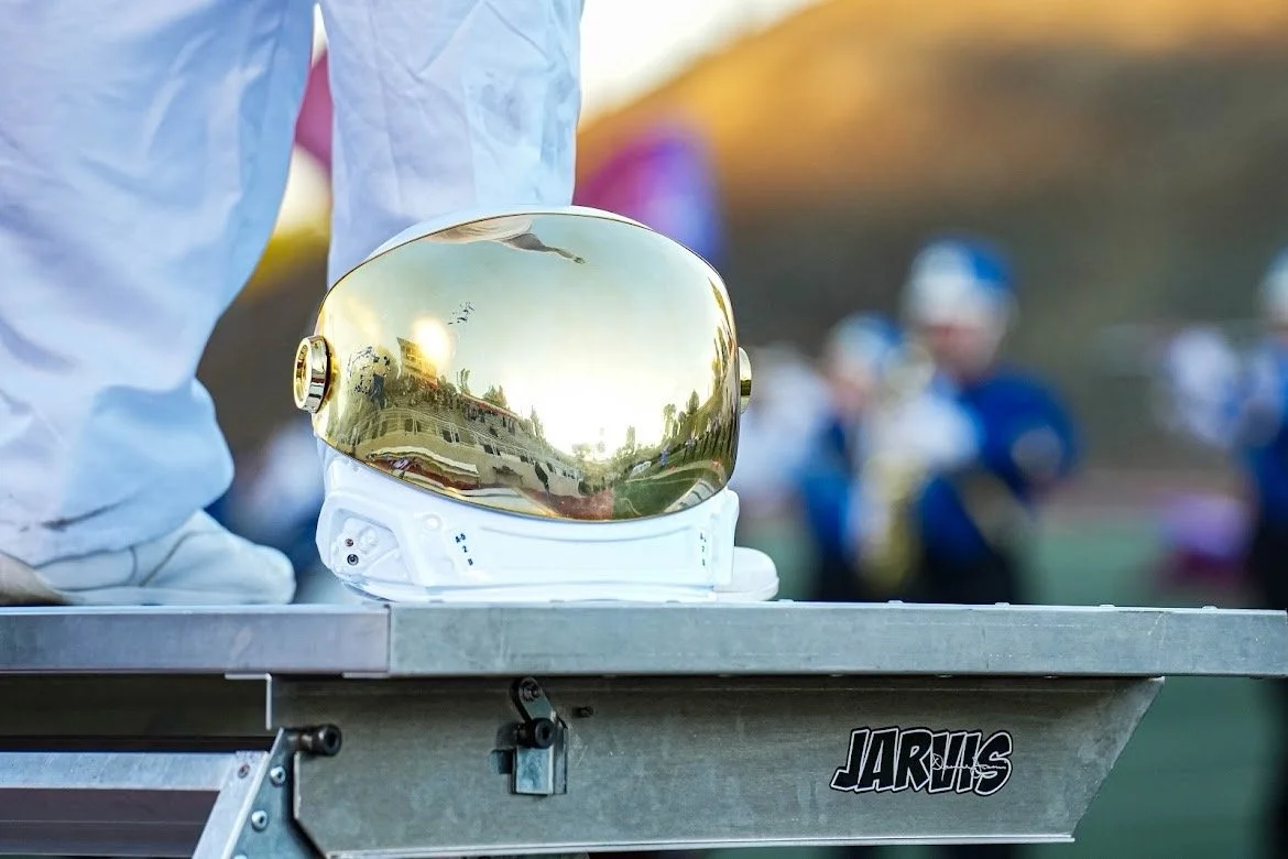 A shiny reflective gold astronaut helmet resting on a white base on a metal table with a background of blurred kids in blue sports uniforms.