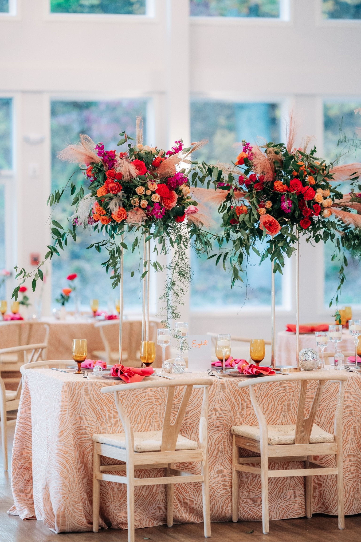 Elegant wedding reception table setup with vibrant floral centerpieces, peach tablecloth, wooden chairs, and glassware.