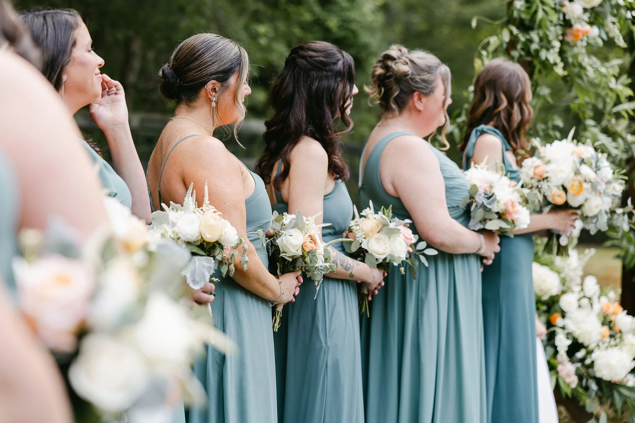 Bridesmaids in green dresses holding bouquets during a wedding ceremony outdoors.