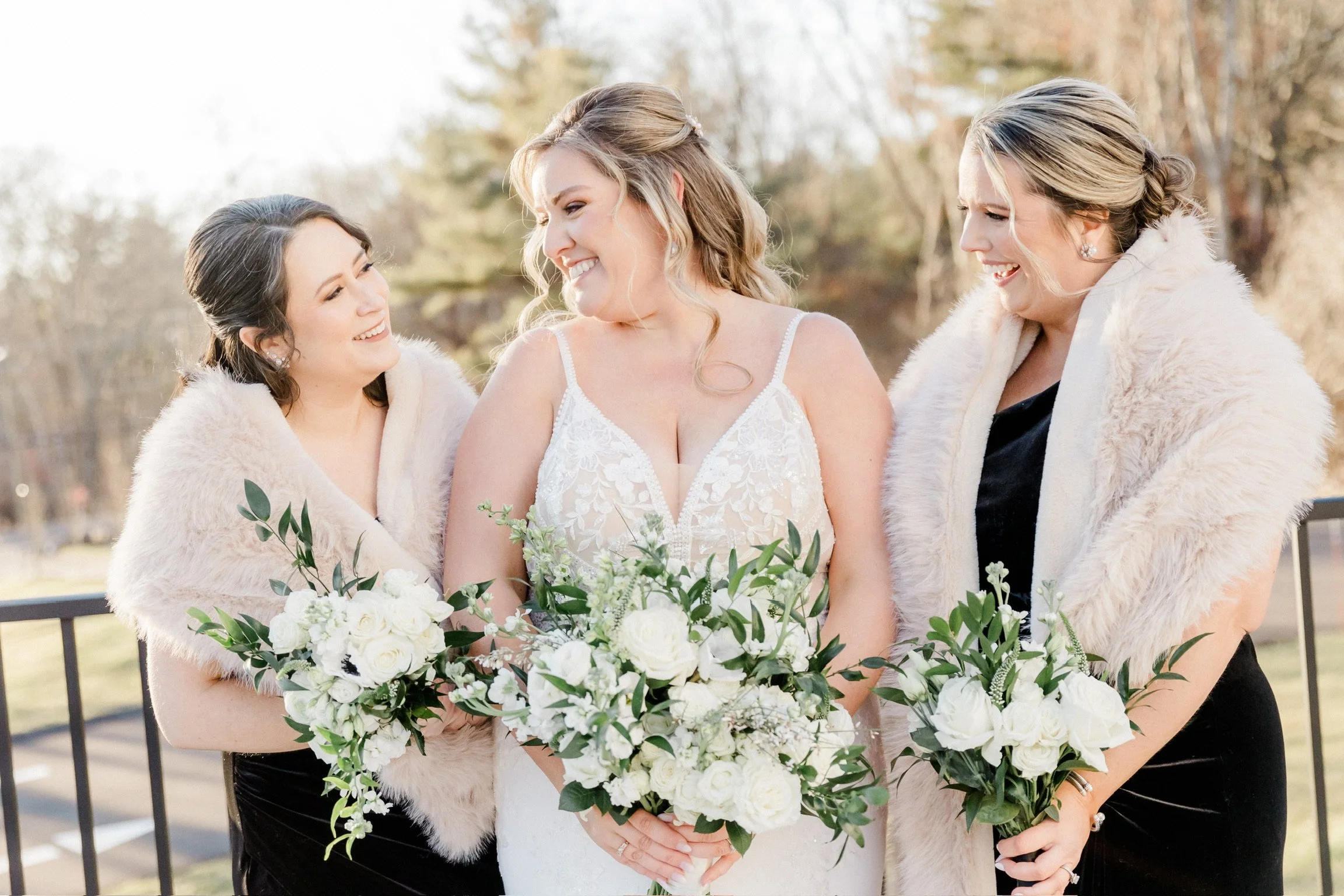 Bride and bridesmaids smiling outdoors, holding white and green floral bouquets, wearing formal dresses and fur stoles.