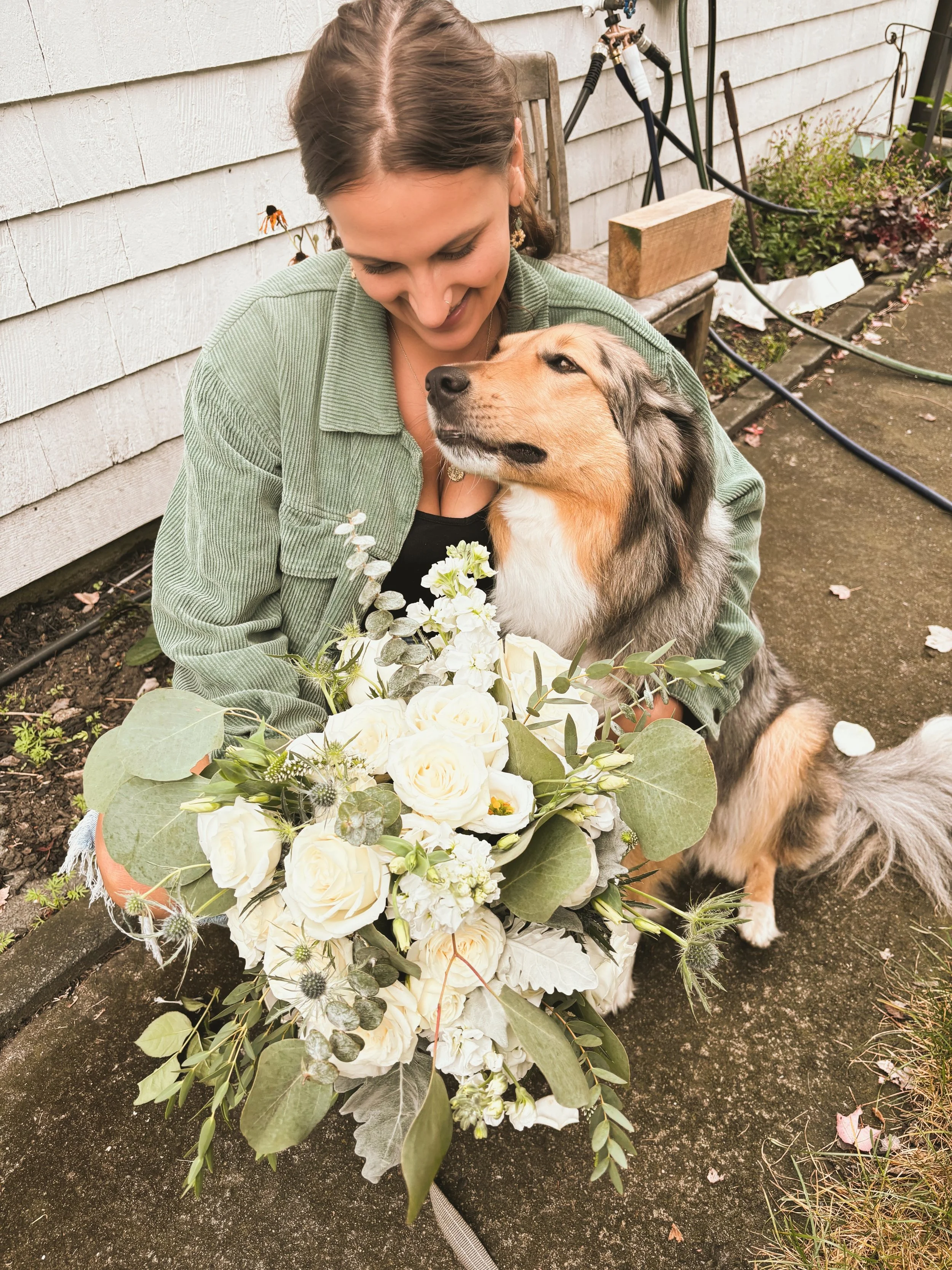 A woman smiling at a dog while holding a large bouquet of white flowers with green leaves, sitting on a concrete path next to a house.