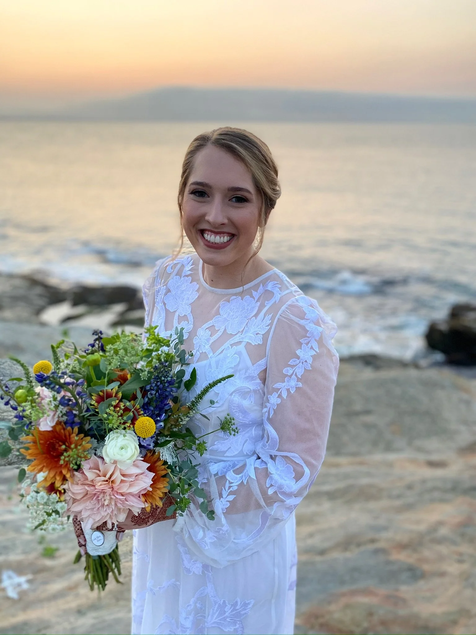 A smiling woman in a white lace dress holding a colorful bouquet of flowers stands by the ocean at sunset.