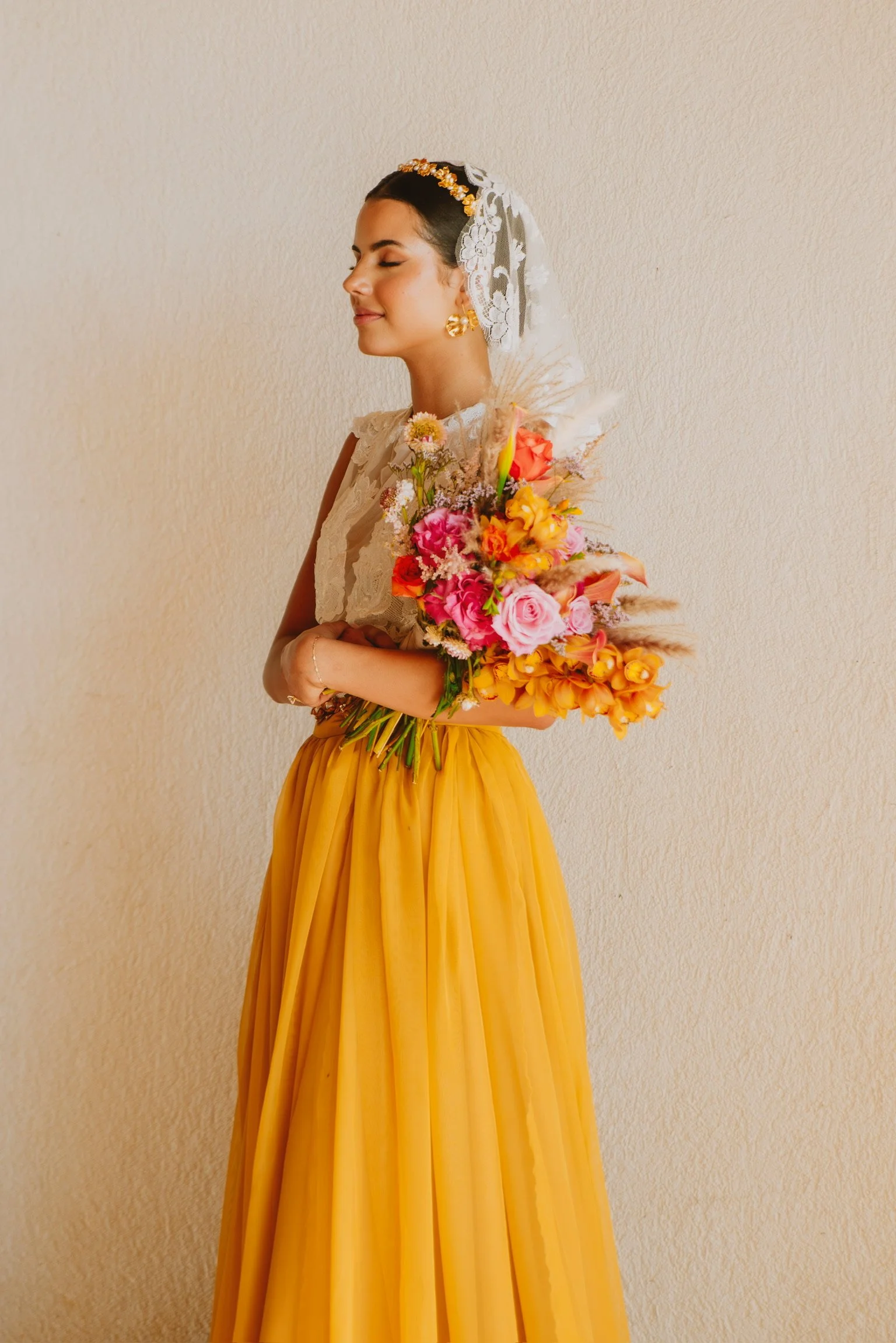 A woman in a yellow skirt, white lace top, and veil holding a vibrant bouquet of colorful flowers against a plain background.