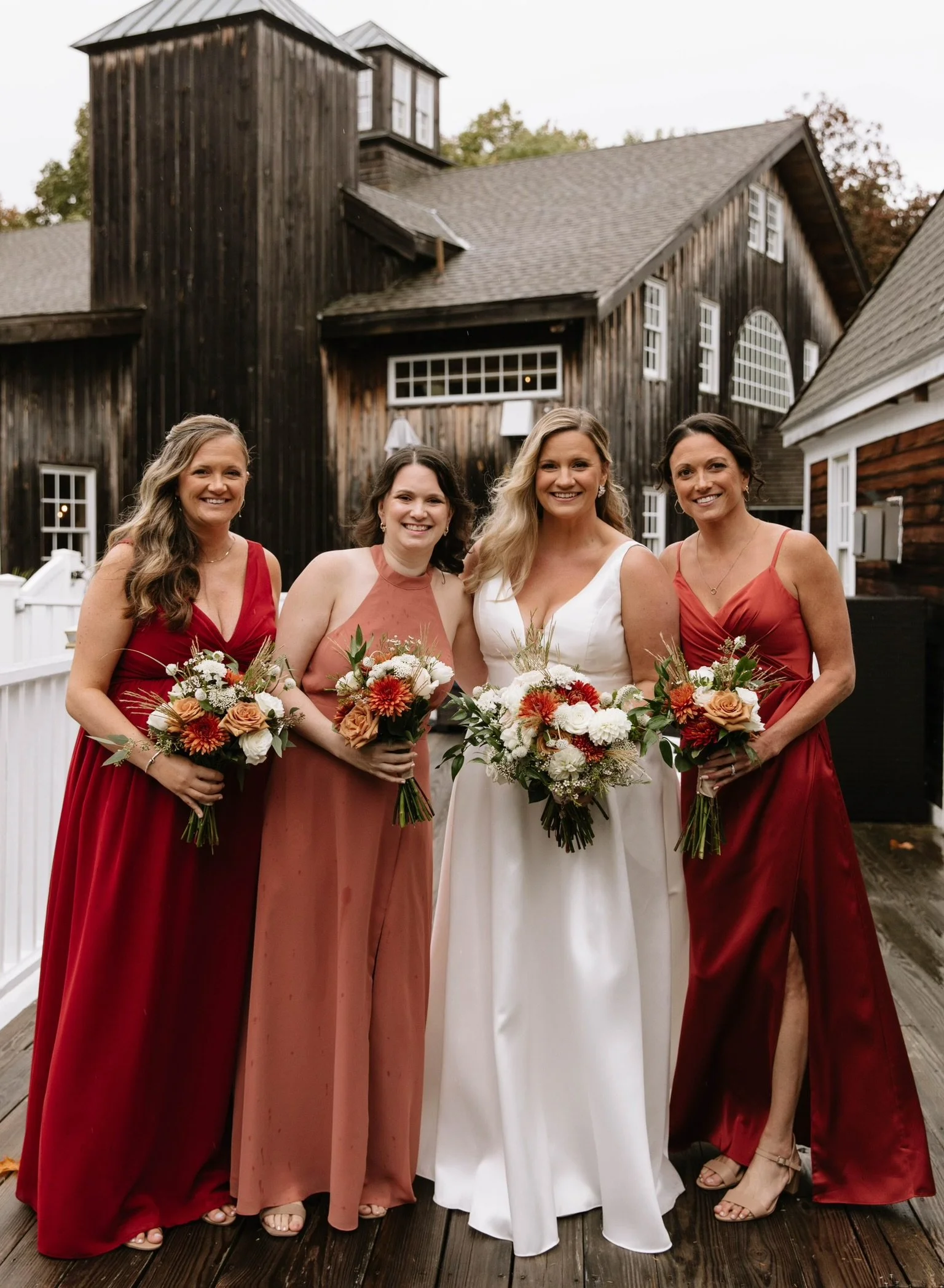A bride in a white wedding dress poses with three bridesmaids wearing red and pink dresses, each holding bouquets of flowers, in front of a rustic wooden building.