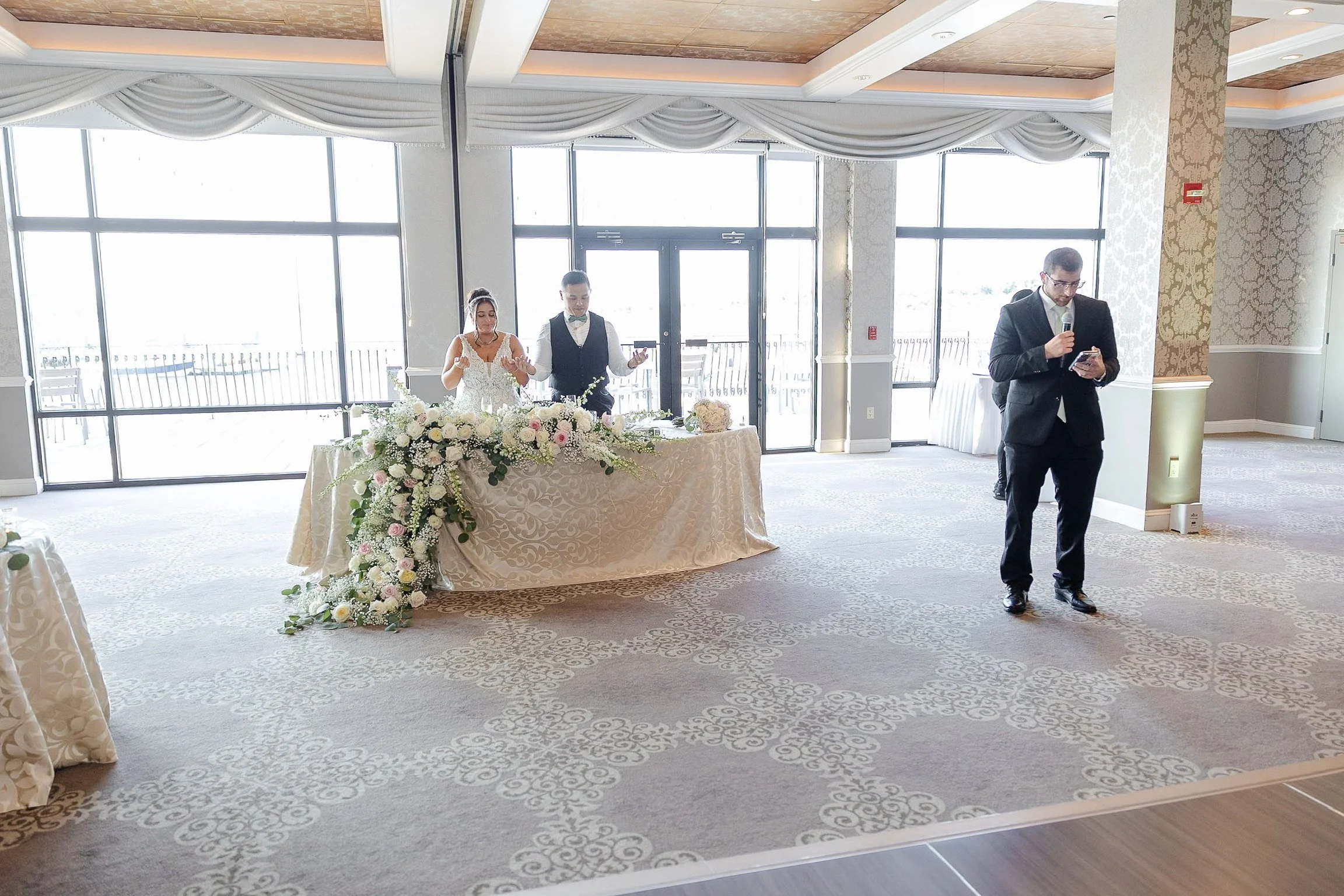 Bride and groom at a decorated wedding reception table with a man giving a speech in a formal venue.
