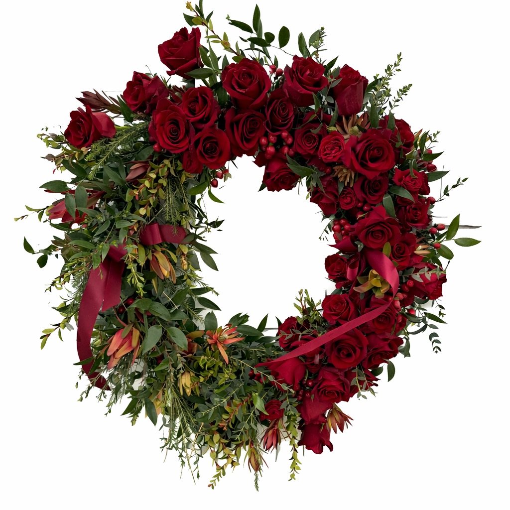 A heart-shaped floral wreath made of red roses, green leaves, and small red berries, decorated with a red ribbon.