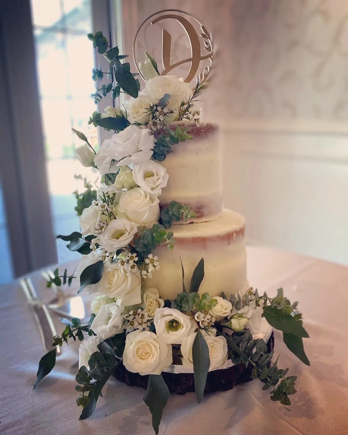 Two-tier naked cake adorned with white roses and greenery topped with a clear acrylic letter ornament.