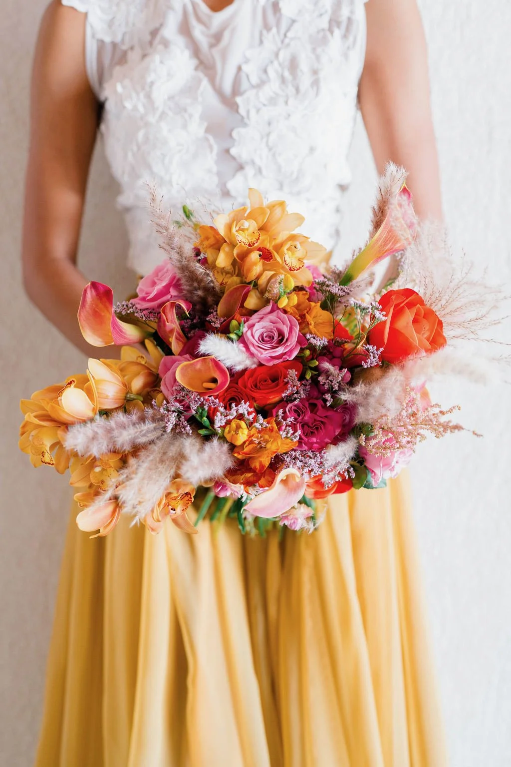 Woman holding a colorful bouquet of flowers, including roses, calla lilies, and pampas grass, wearing a white textured top and a yellow skirt.