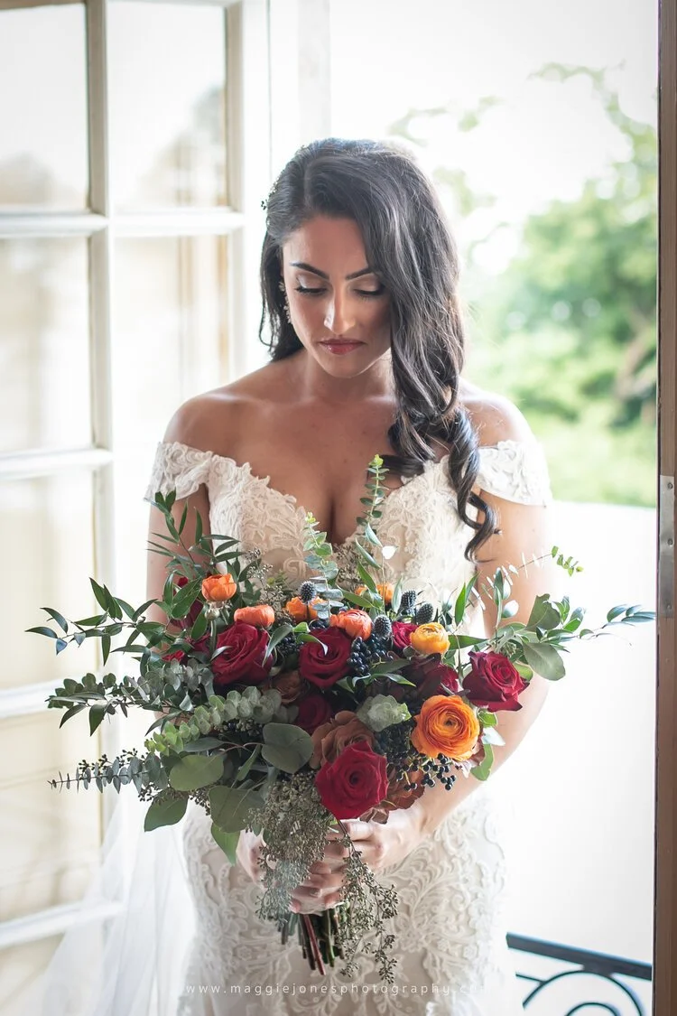 Bride in lace wedding dress holding a bouquet of red and orange flowers, standing by a window.
