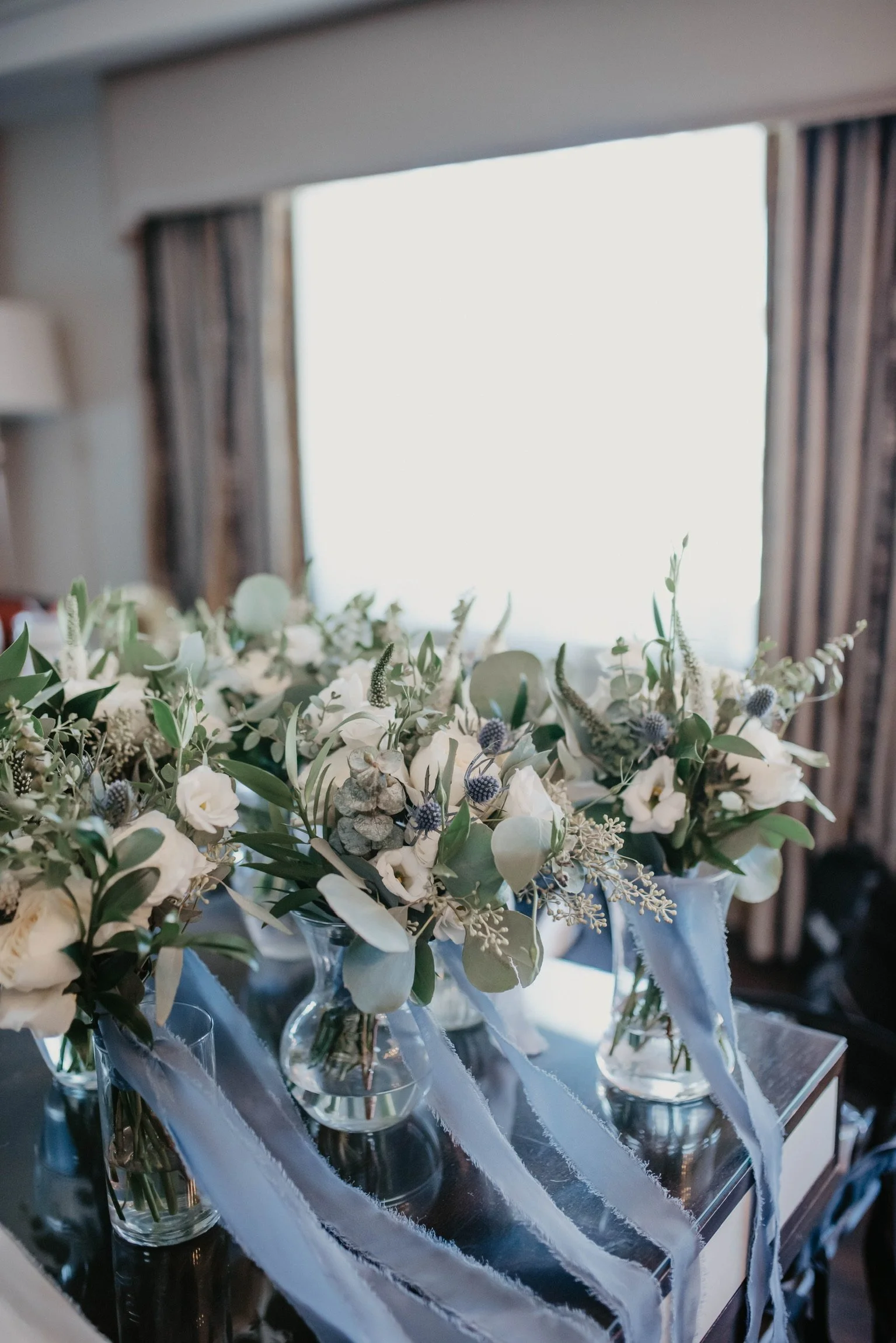 Bouquets of white roses and greenery in glass vases with blue ribbons on a table.
