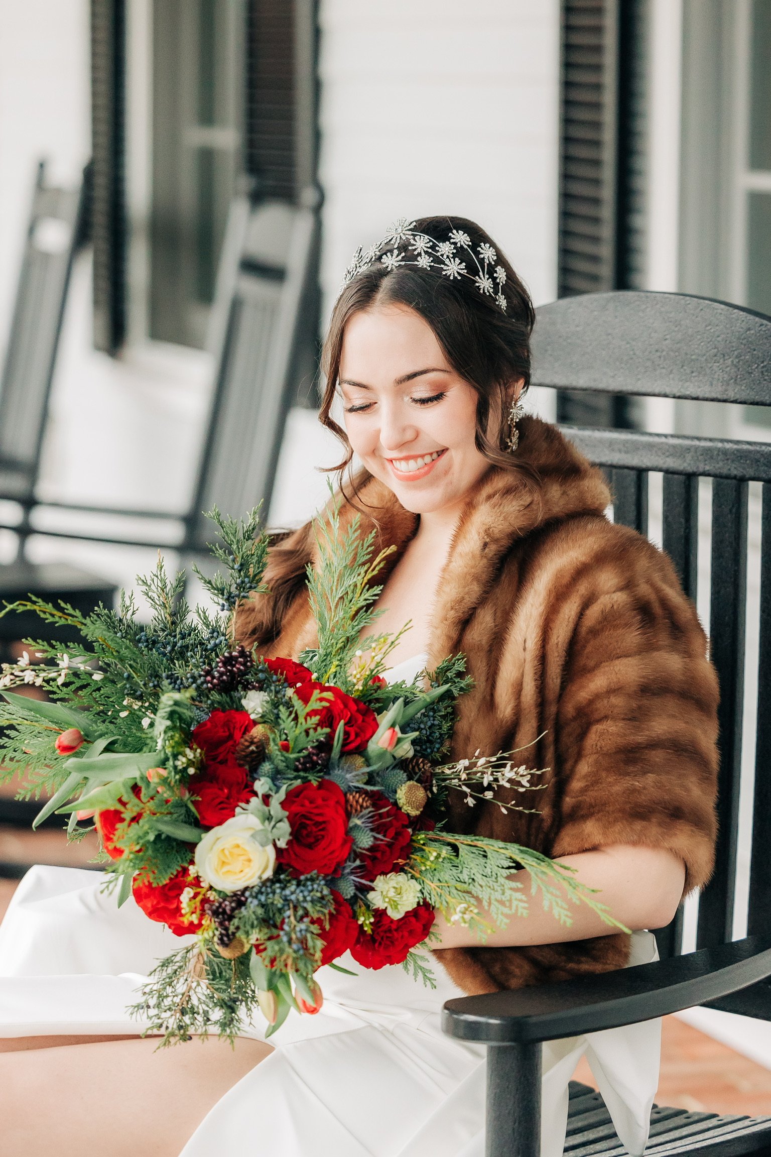 Bride in a fur wrap holding a red and green bouquet while sitting on a porch.