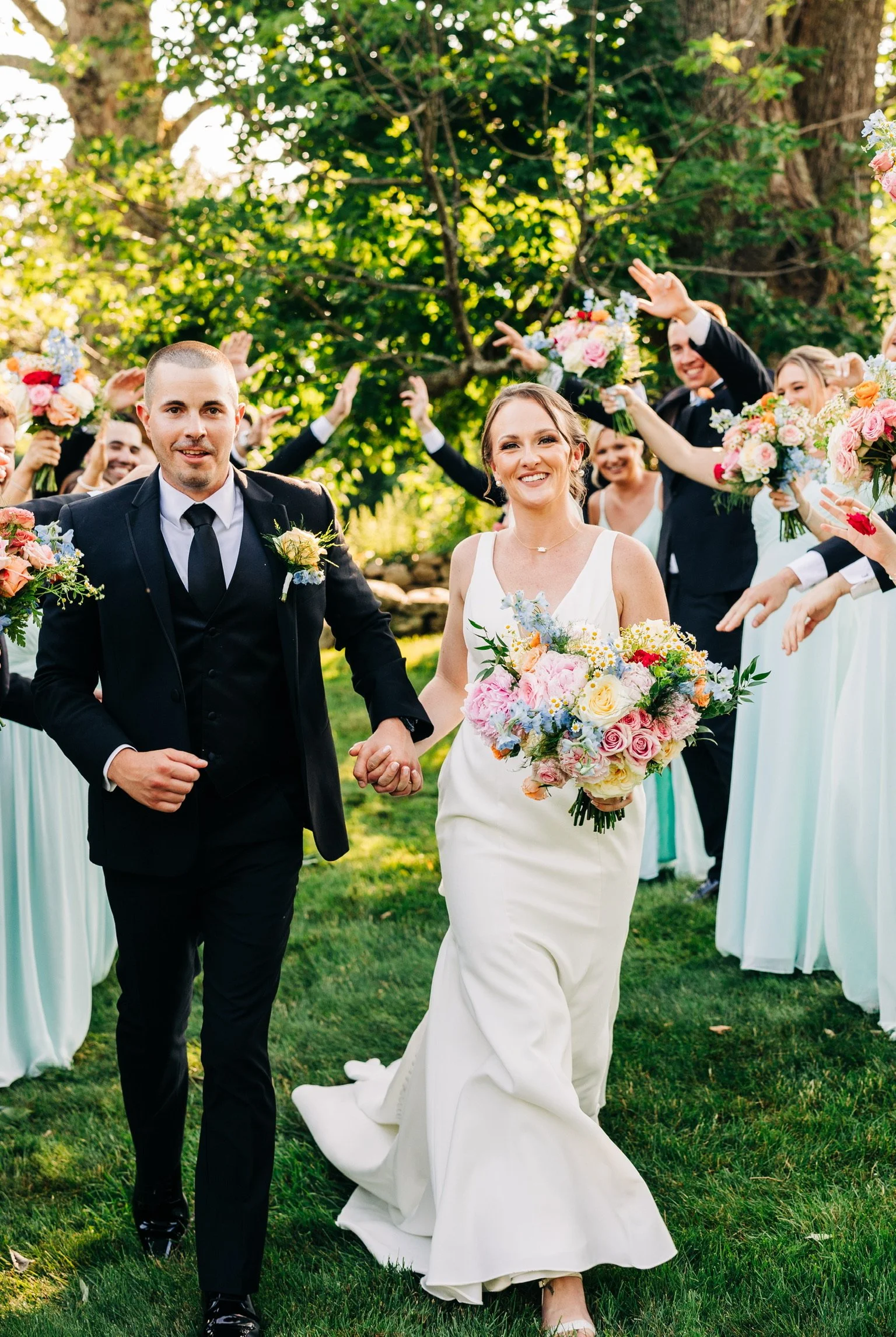 A smiling bride and groom walk hand in hand, surrounded by a joyful bridal party holding colorful bouquets, in an outdoor, garden setting.