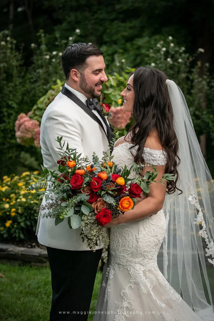 Bride and groom in wedding attire, outdoors, holding a bouquet of red and orange flowers.