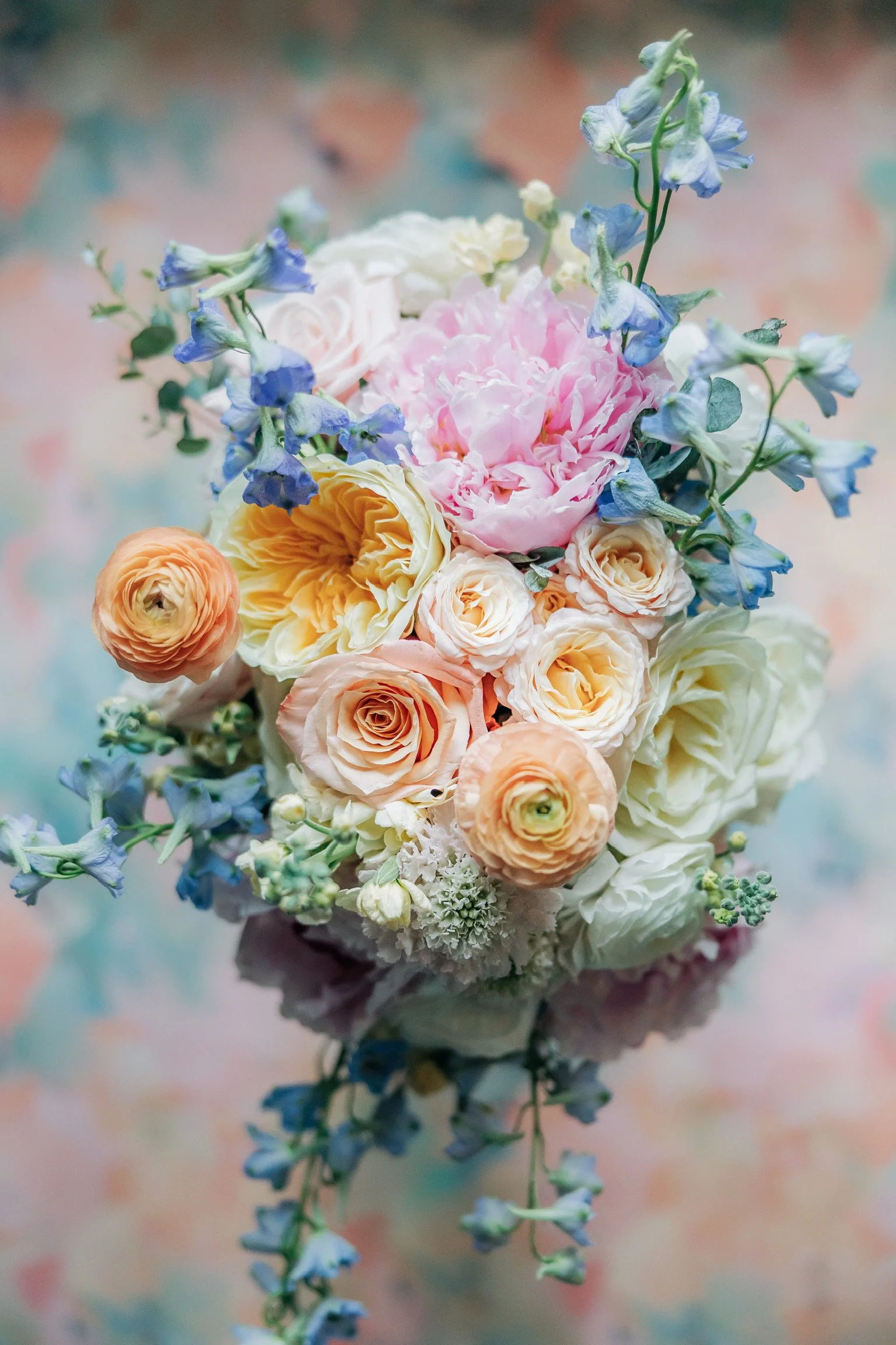 A colorful flower bouquet with roses, peonies, and delphiniums against a soft pastel background.