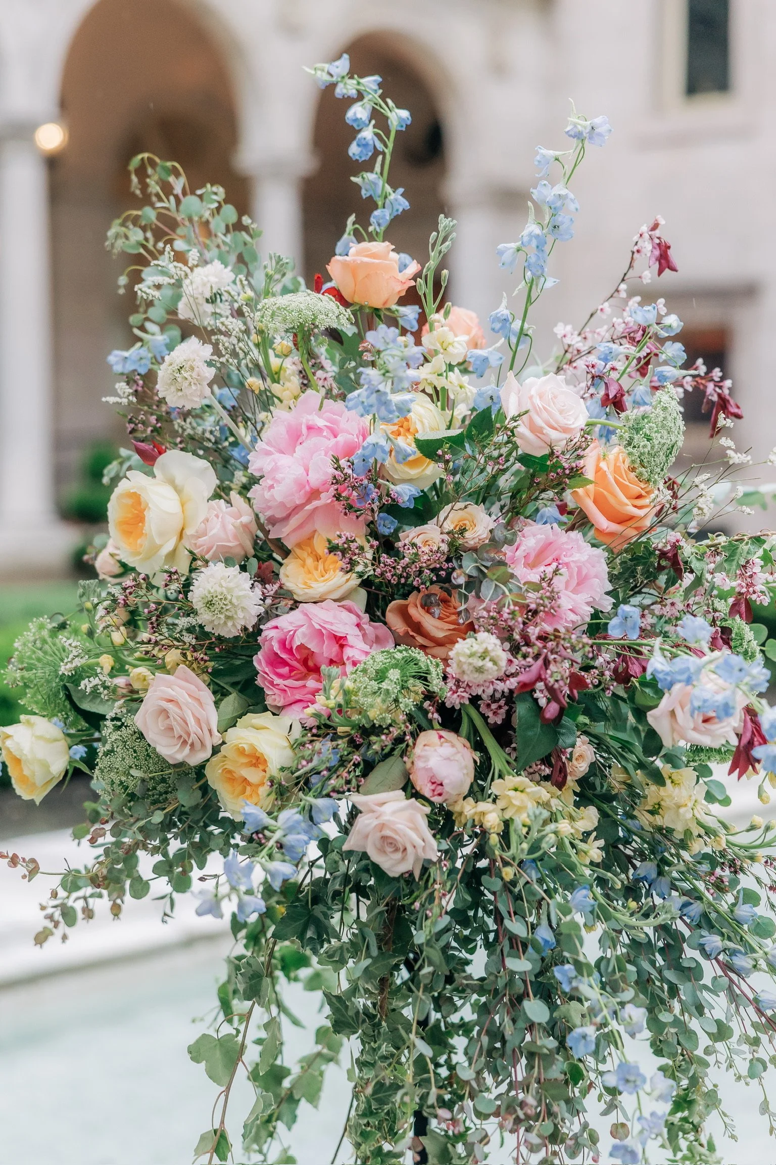 Colorful floral arrangement with roses, peonies, and greenery in a courtyard setting.
