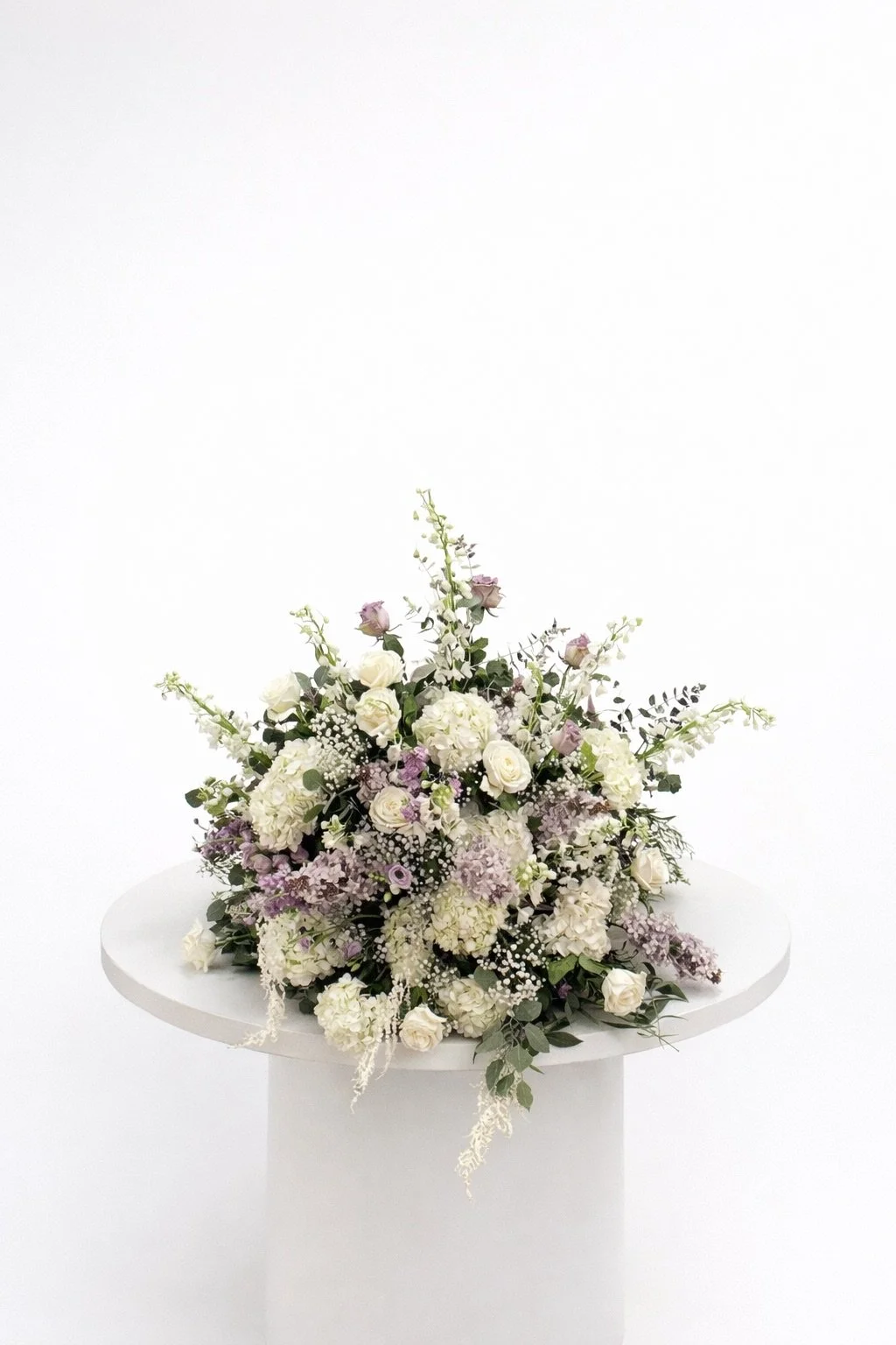 Elegant floral arrangement with white and light purple flowers on a white pedestal against a plain white background.