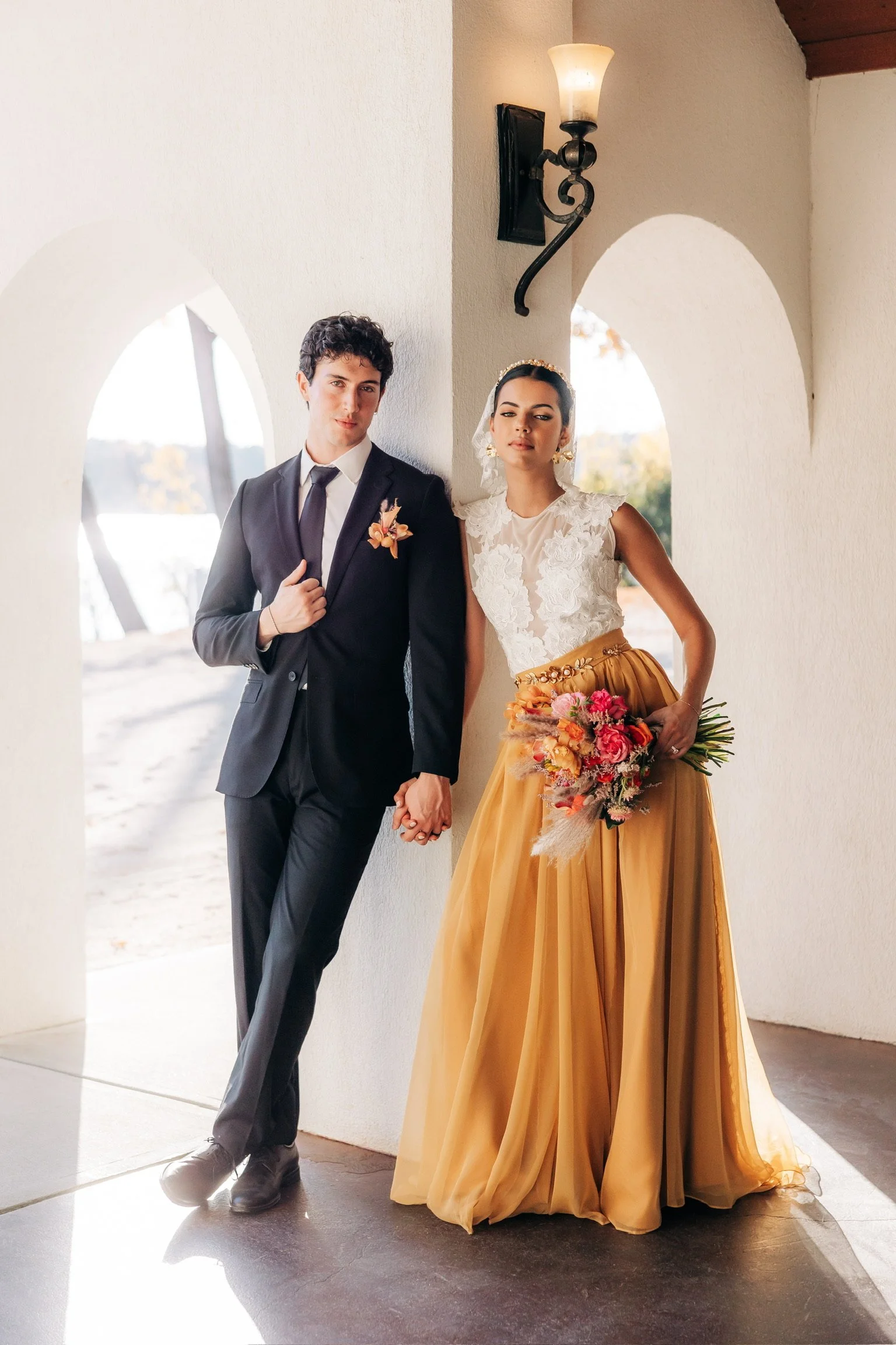 A couple holding hands, dressed in formal attire, with the woman wearing a lace top and mustard-colored skirt, holding a bouquet, and the man in a suit with a boutonniere, standing under an archway.