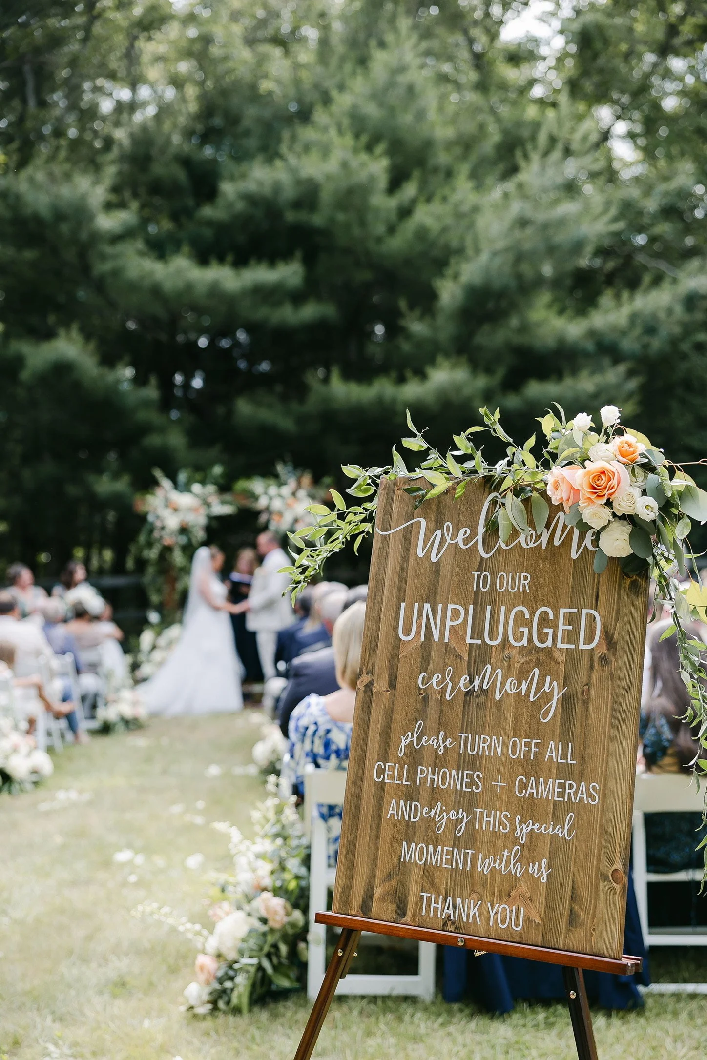 Outdoor wedding ceremony with a wooden sign stating "Welcome to our unplugged ceremony, please turn off all cell phones + cameras" in front of guests and a couple getting married under a floral arch.