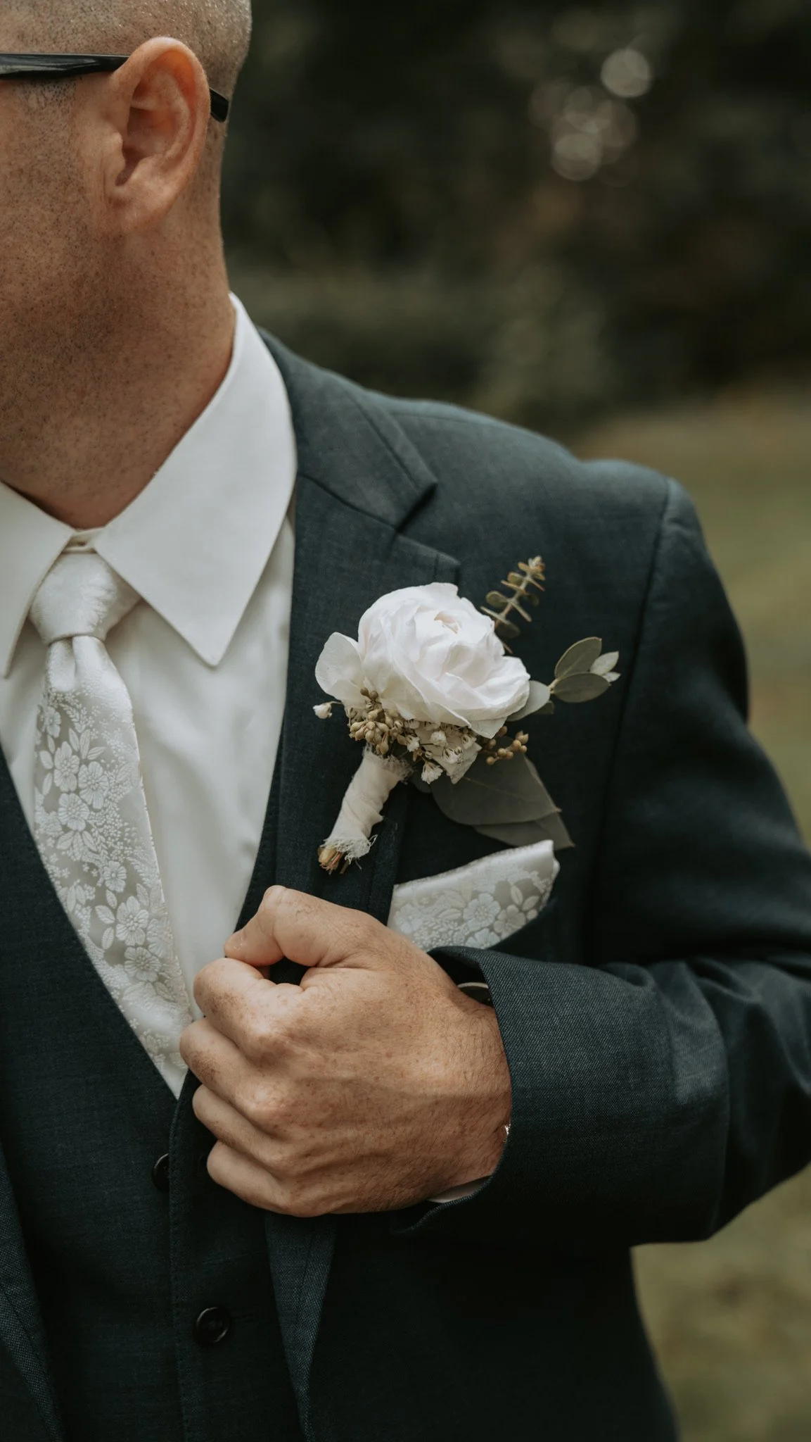 Man in dark suit with white rose boutonniere, white floral tie, holding lapel, blurred background.