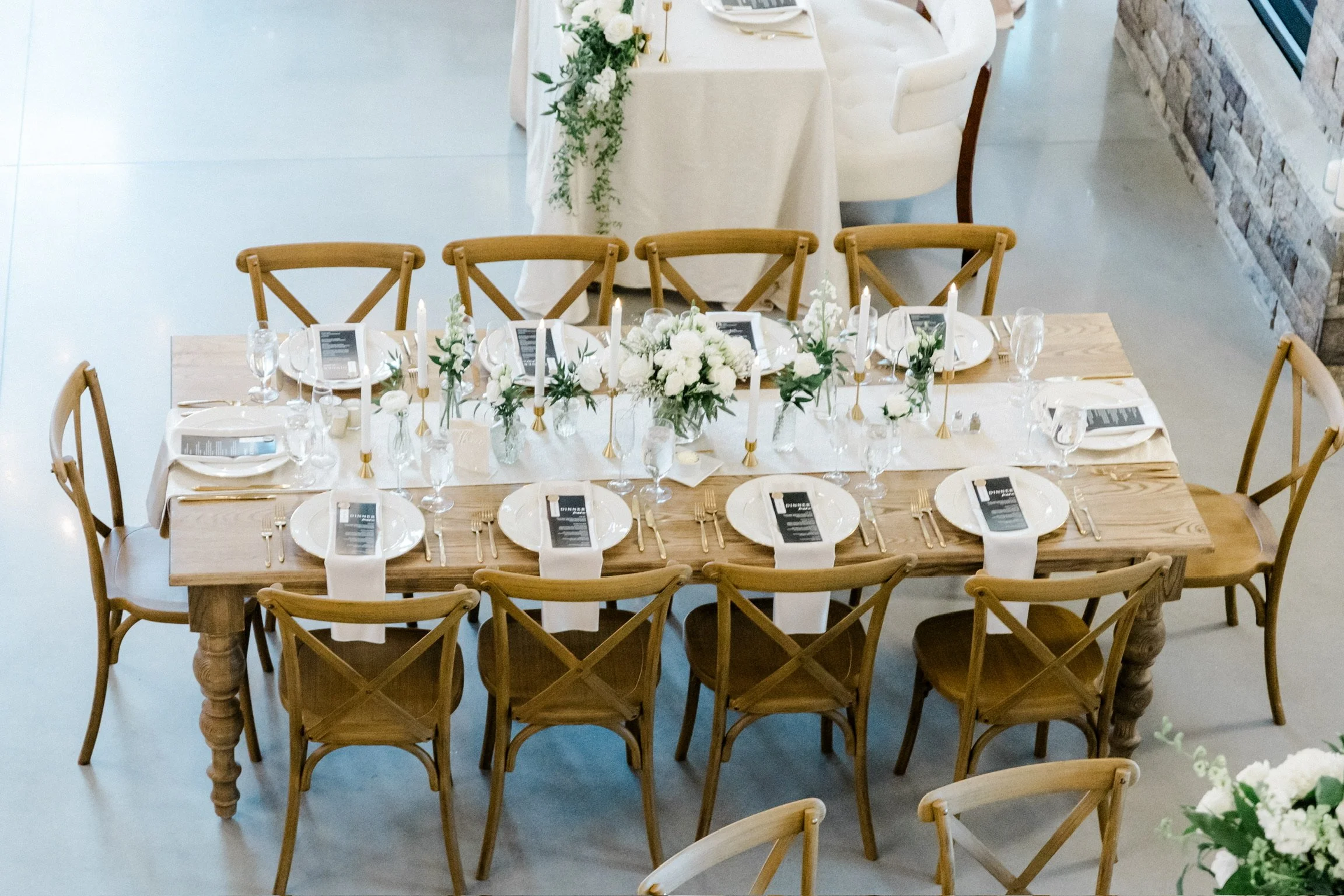 Elegant dining table setup for a wedding with wooden chairs, white plates, menus, candles, and floral centerpieces.